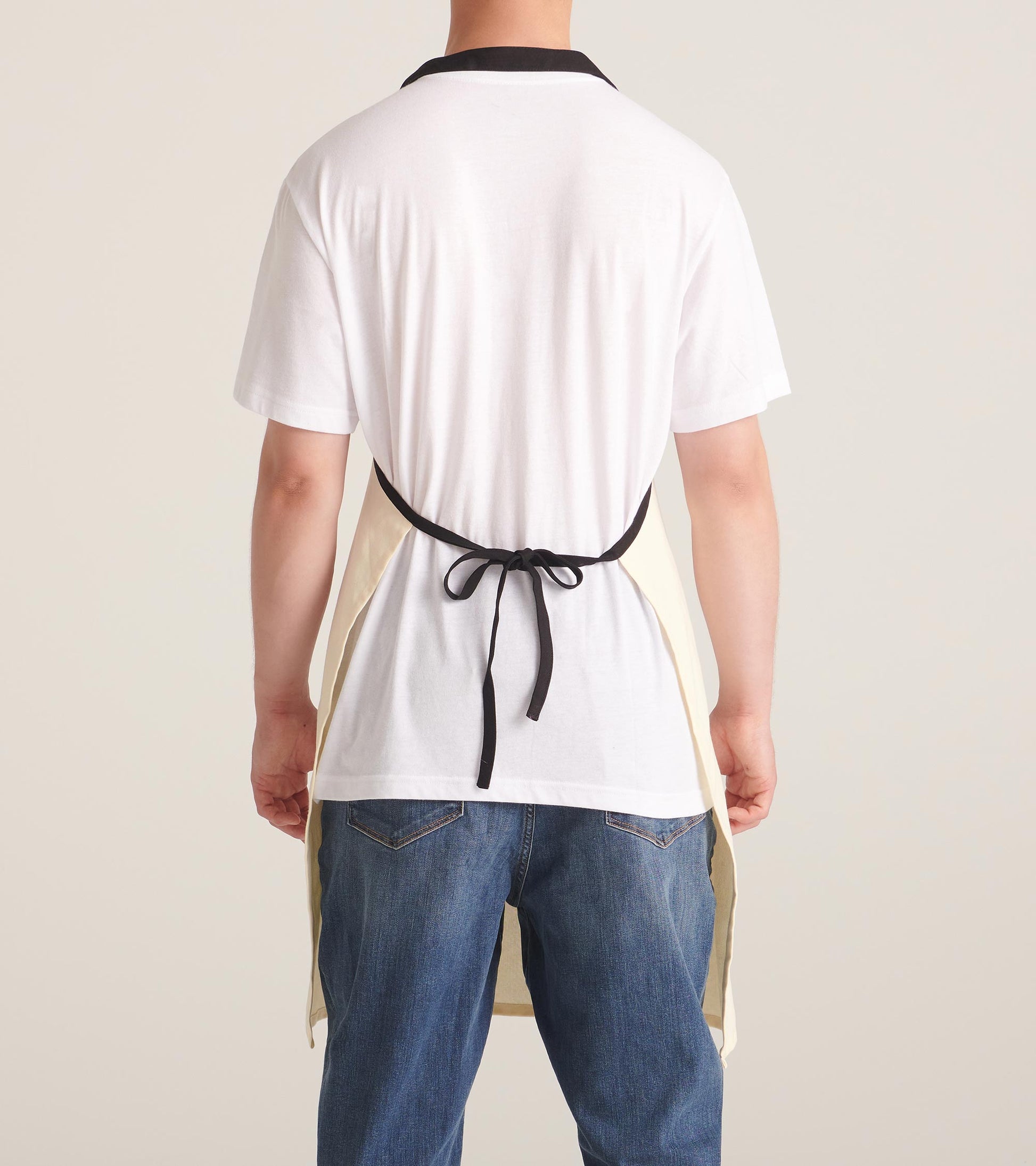 Back view of a cream apron with a black collar, tied at the waist, worn over a white shirt and blue jeans.