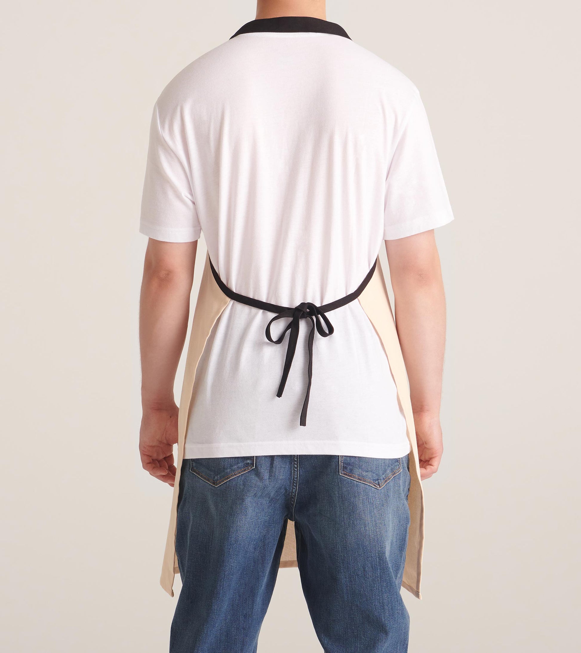 Beige apron with black trim, tied at the back, worn over a white shirt and blue jeans.