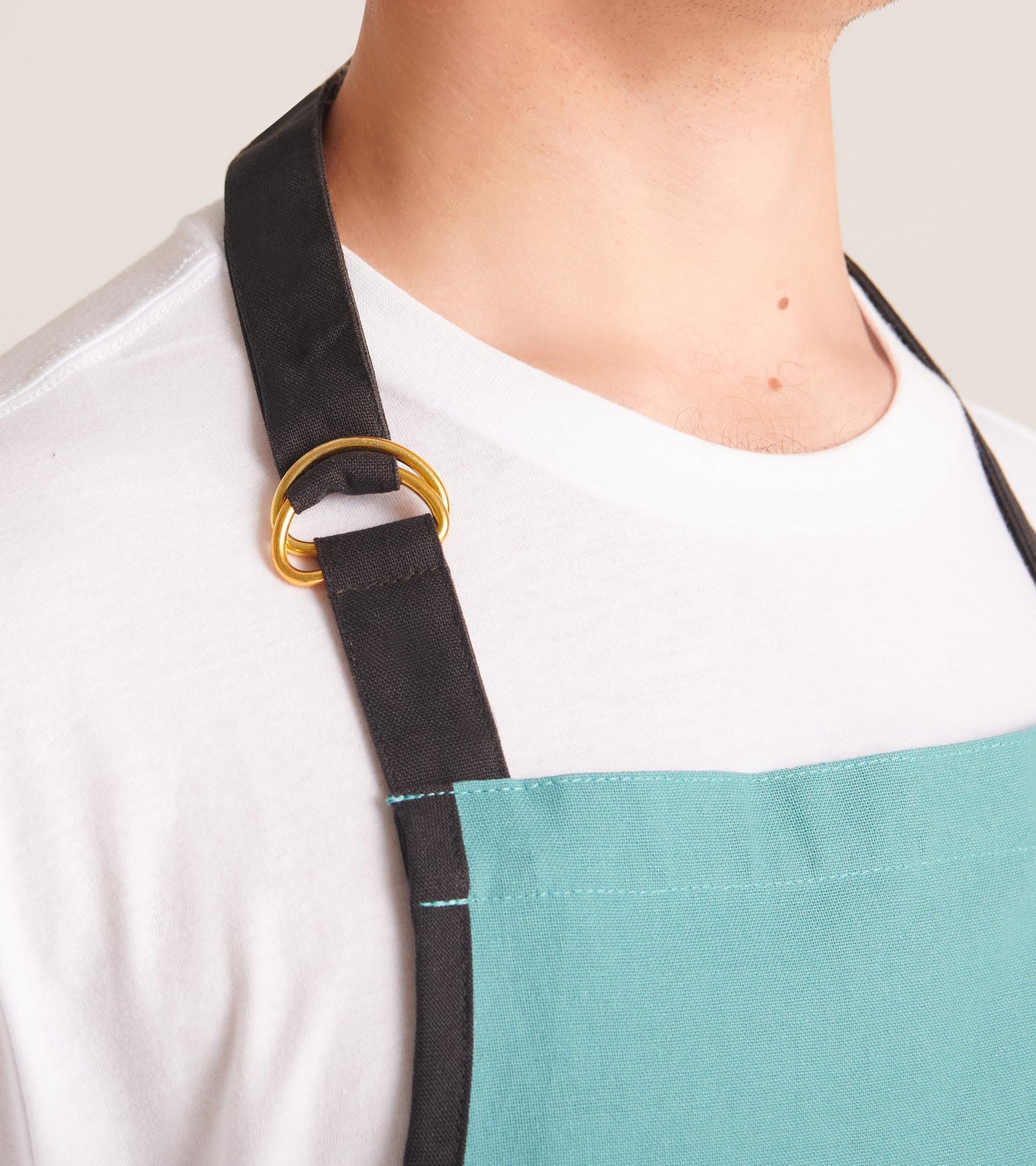Close-up of a white apron with black neck strap, gold ring adjuster, and turquoise front panel