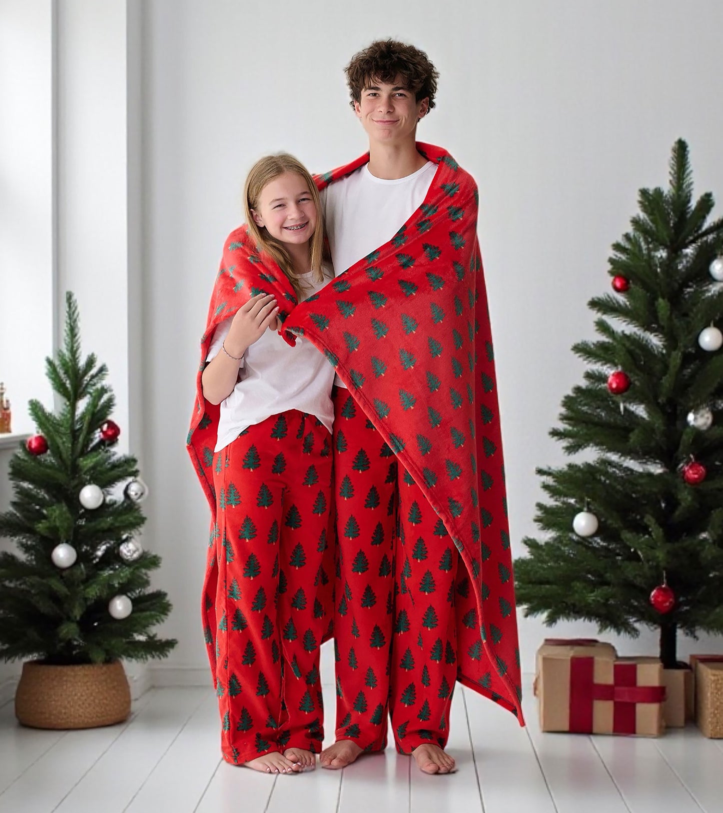 Red fleece blanket with green Christmas tree pattern, shown draped around two children standing indoors.