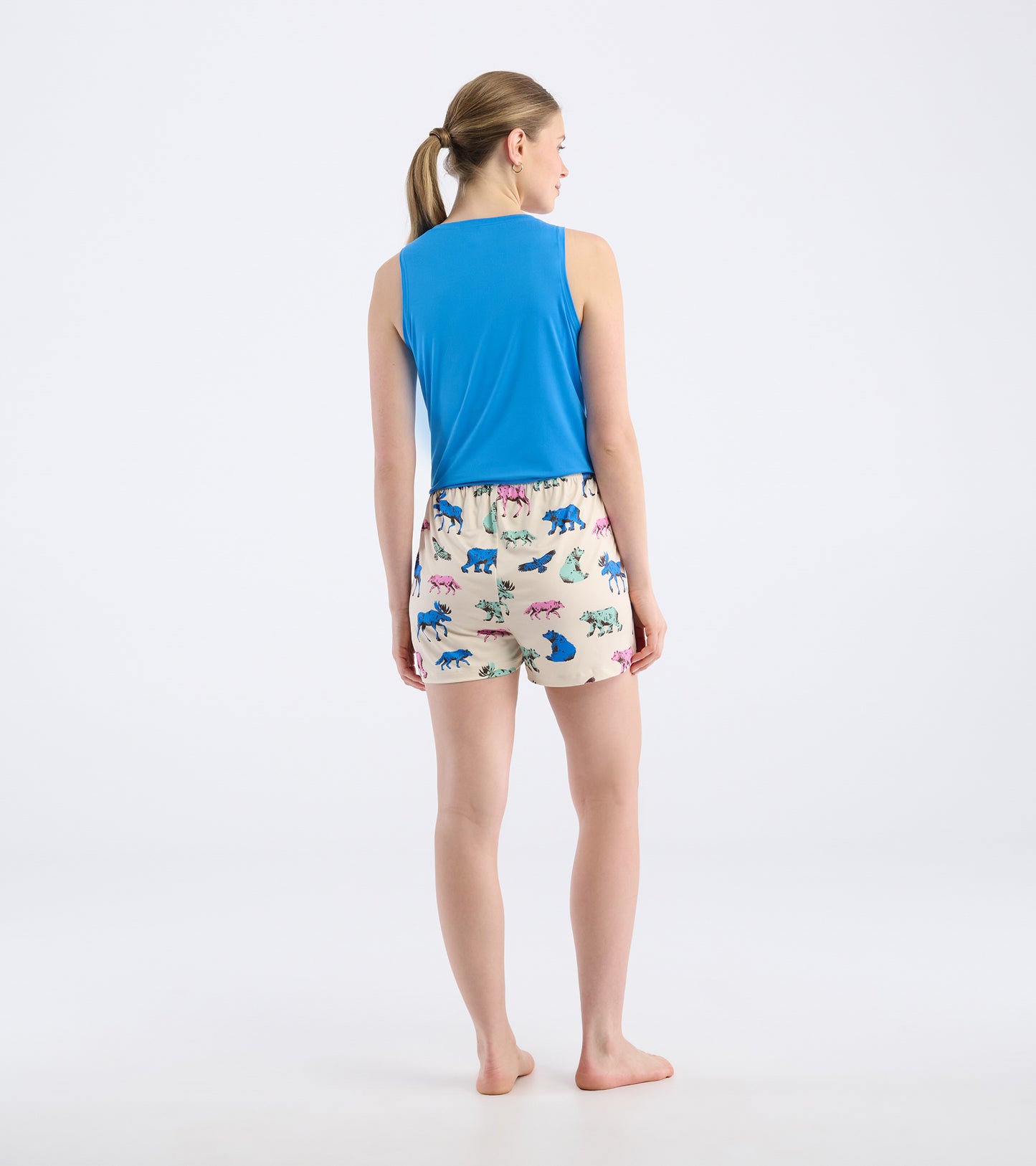 Back view of a woman wearing blue sleeveless top and cream pajama shorts with colorful woodland animal prints.