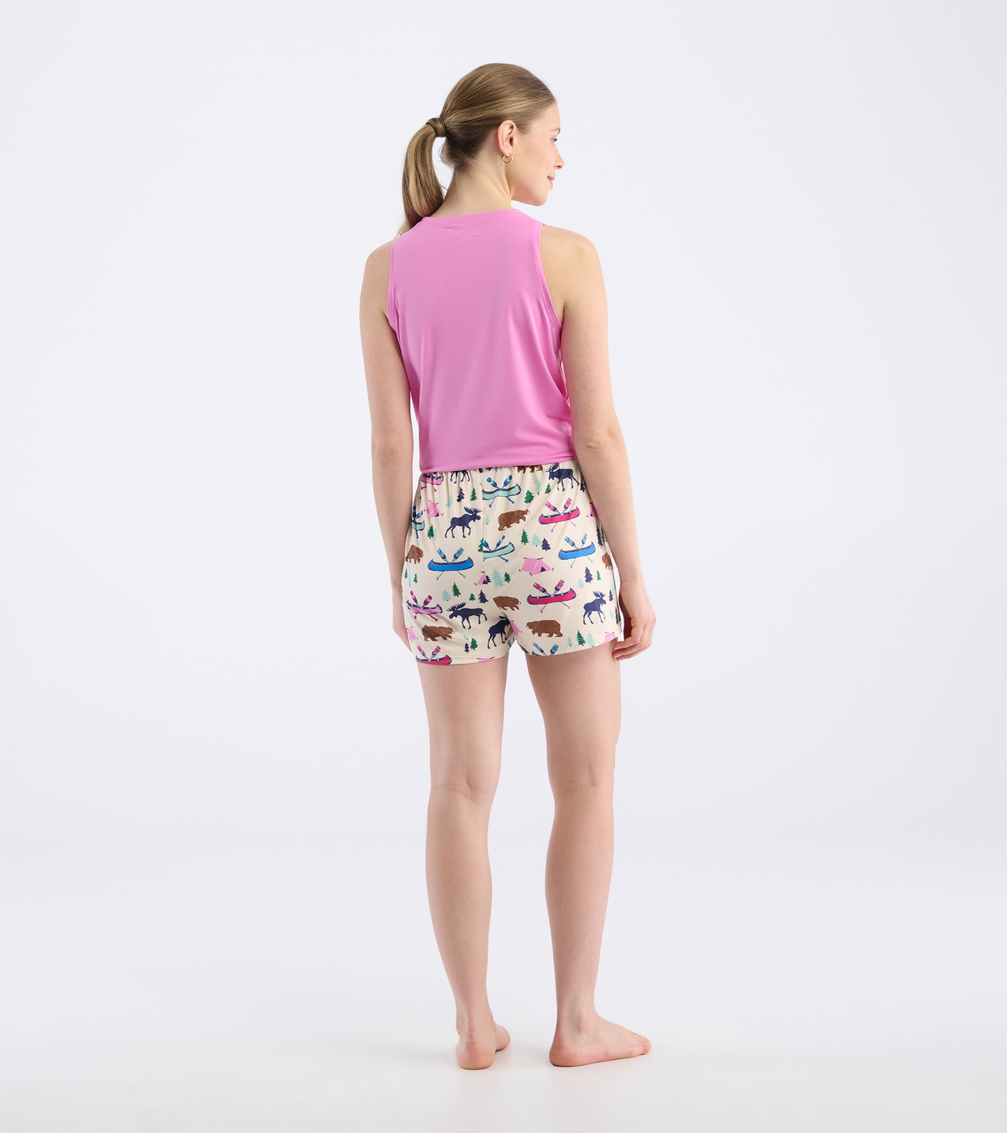 Back view of a woman wearing pink sleeveless top and cream pajama shorts with colorful animal and nature patterns.
