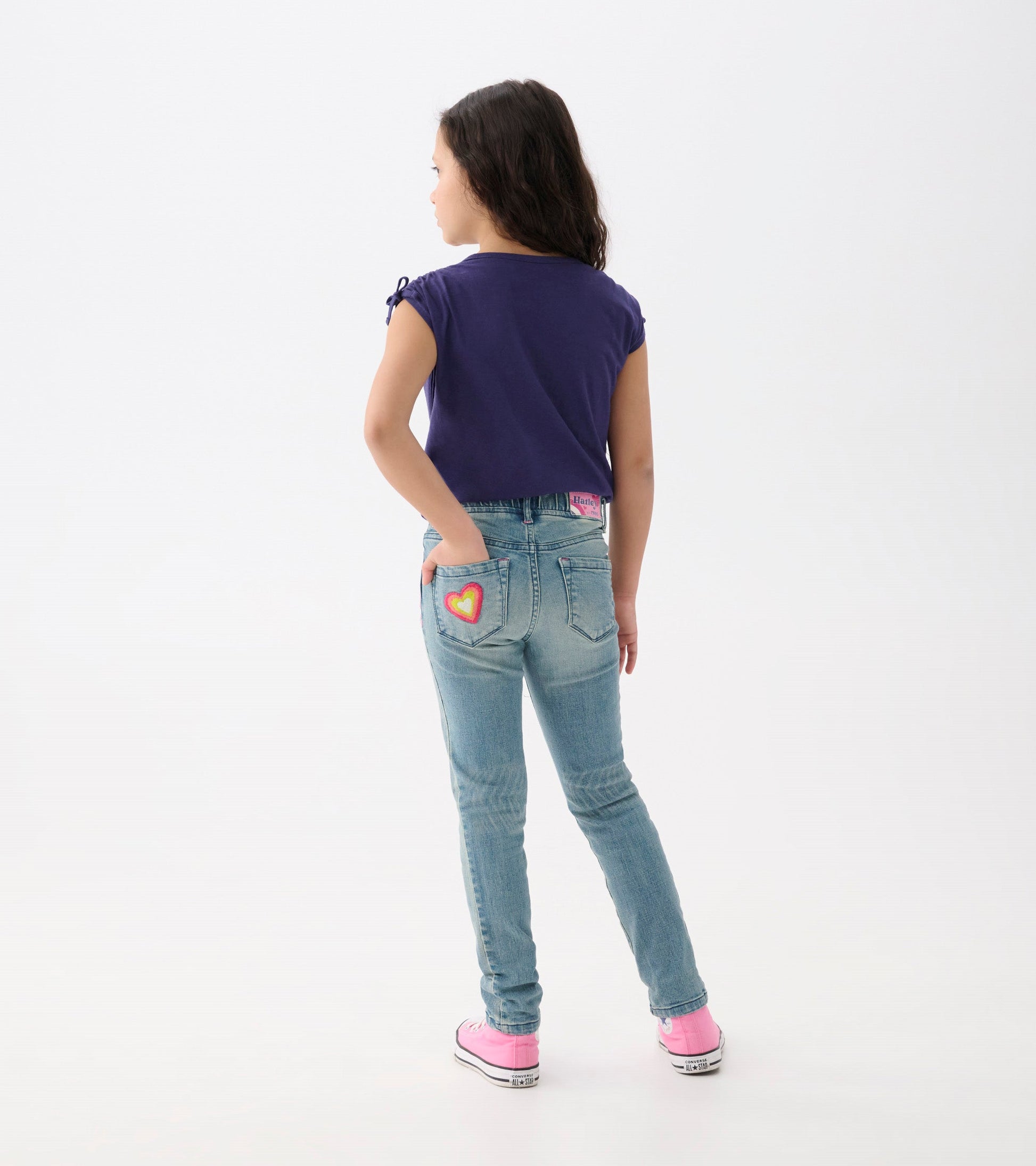 Light blue stretch jeans with colorful heart patch on back pocket, worn by a girl with dark hair, facing away.