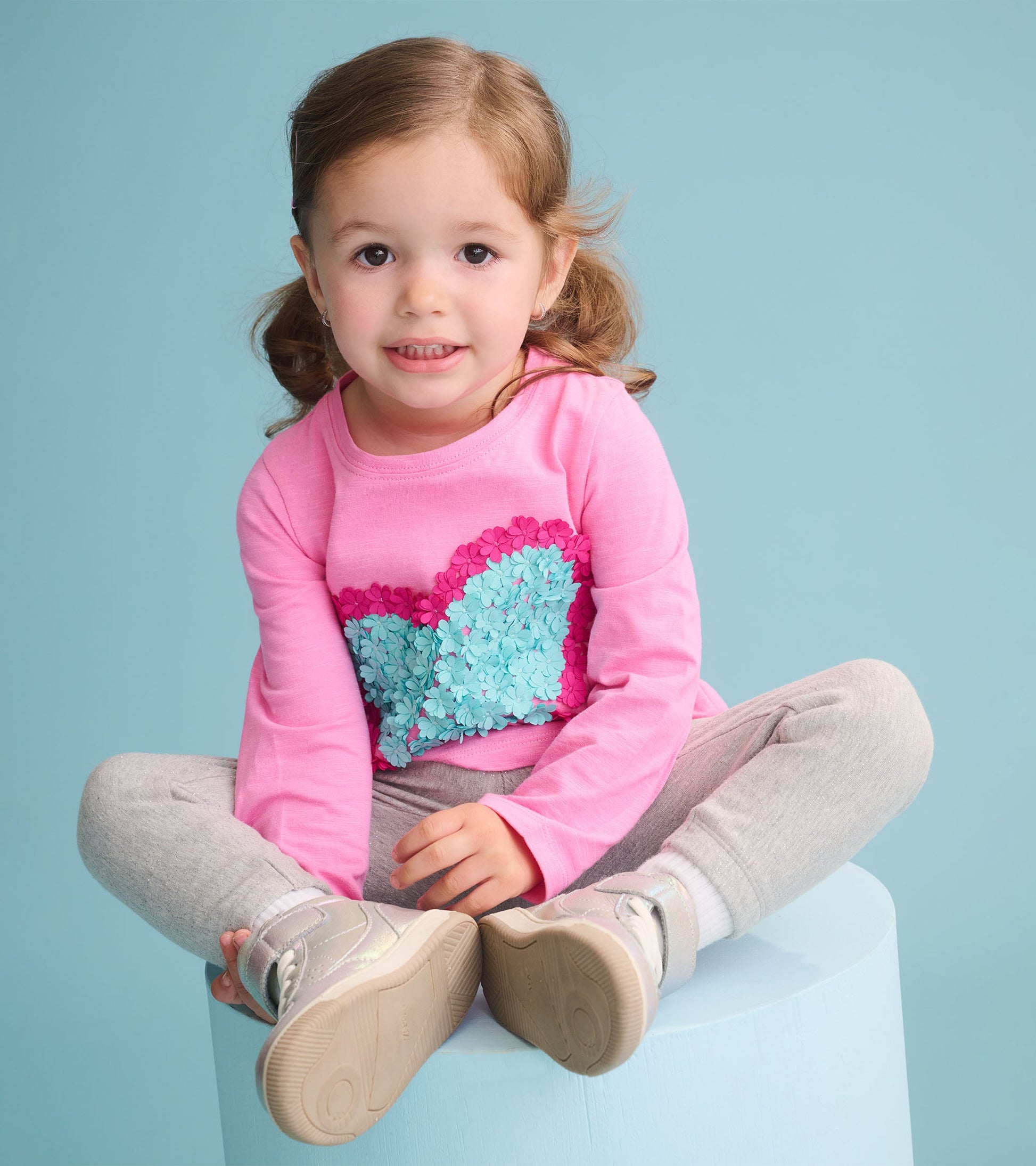 Pink long sleeve t-shirt with a heart design made of blue and pink flower appliqués, paired with gray pants.