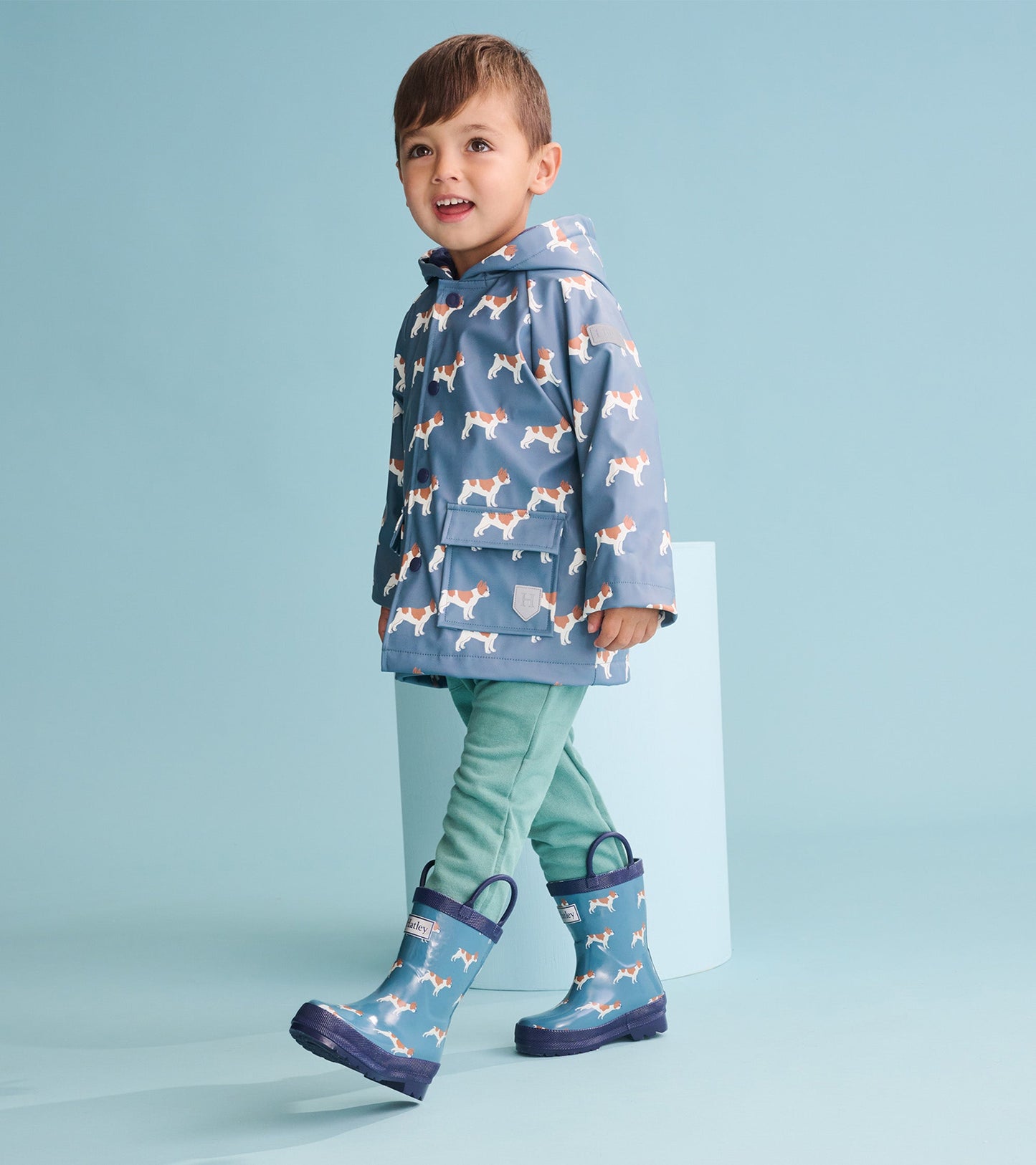 Blue rain jacket with French bulldog print, hood, and front pockets, paired with green pants and matching rain boots.