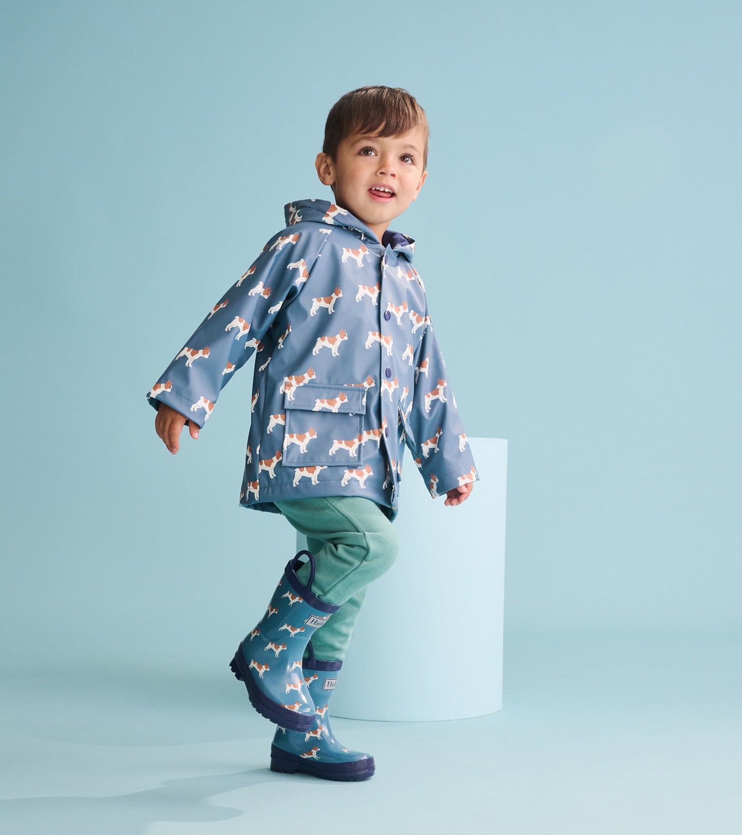 Blue rain jacket with French bulldog pattern, hood, and pockets, worn by a boy with green pants and matching boots.