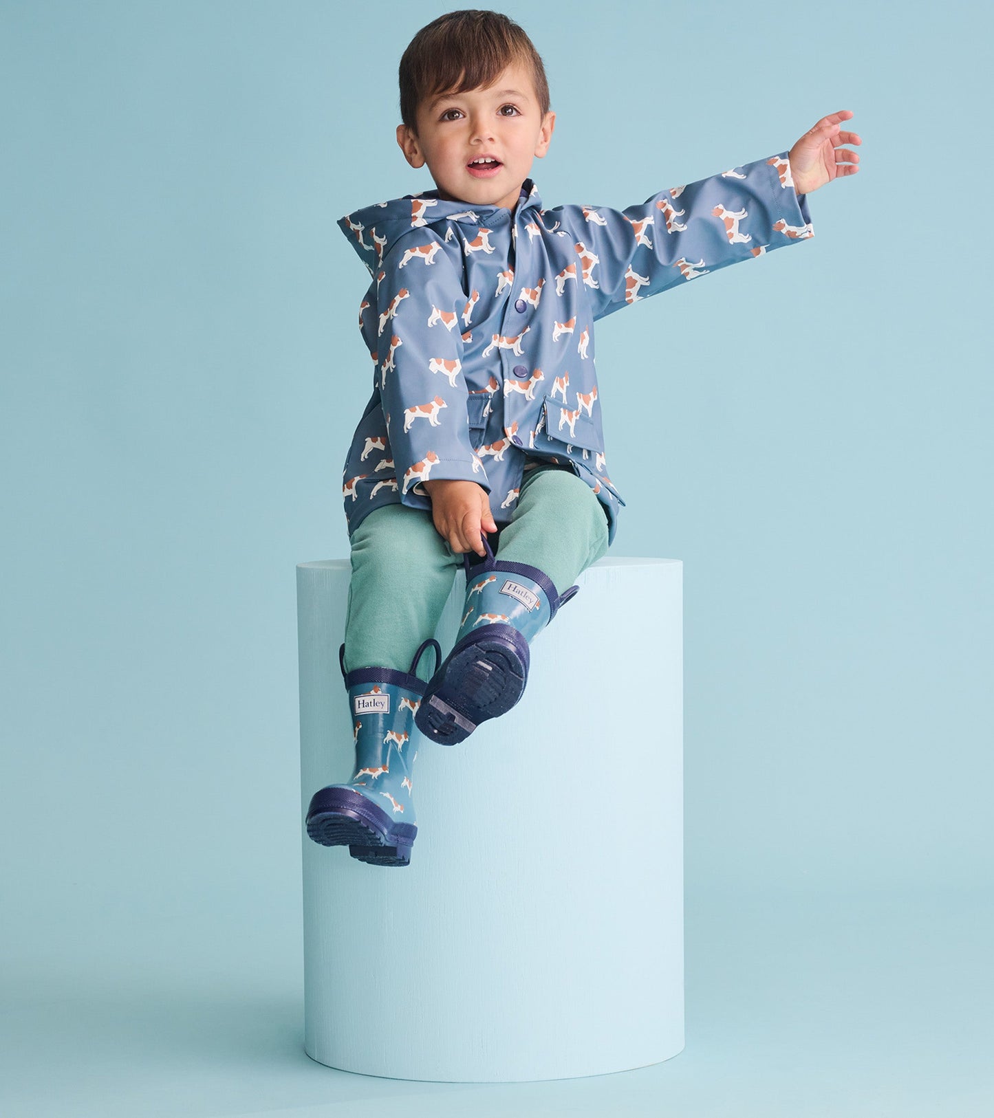 Blue rain jacket with French bulldog pattern, worn by a boy sitting on a pedestal, paired with green pants and rain boots.