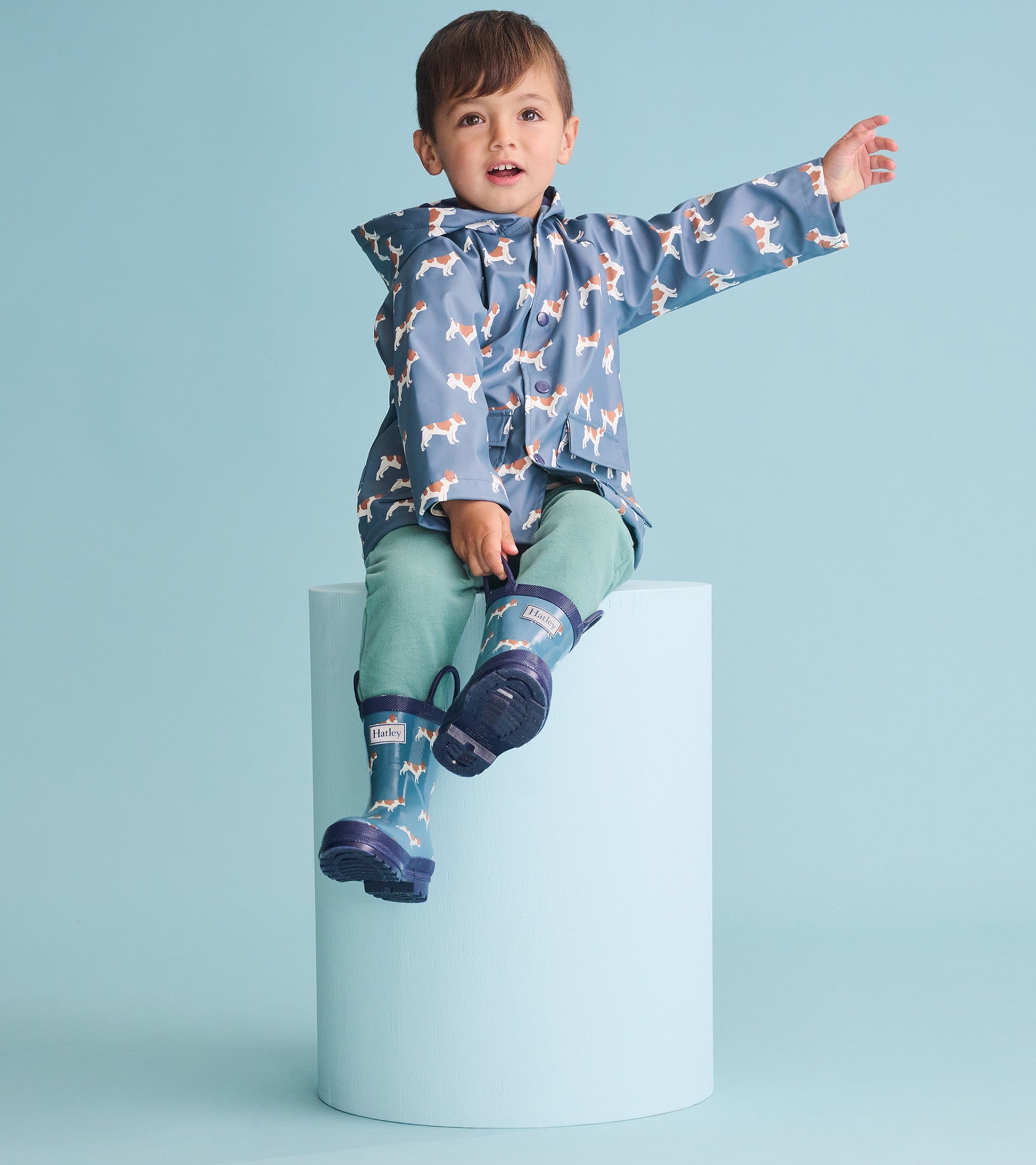 Blue rain jacket with French bulldog pattern, worn by a boy sitting on a pedestal, paired with green pants and rain boots.