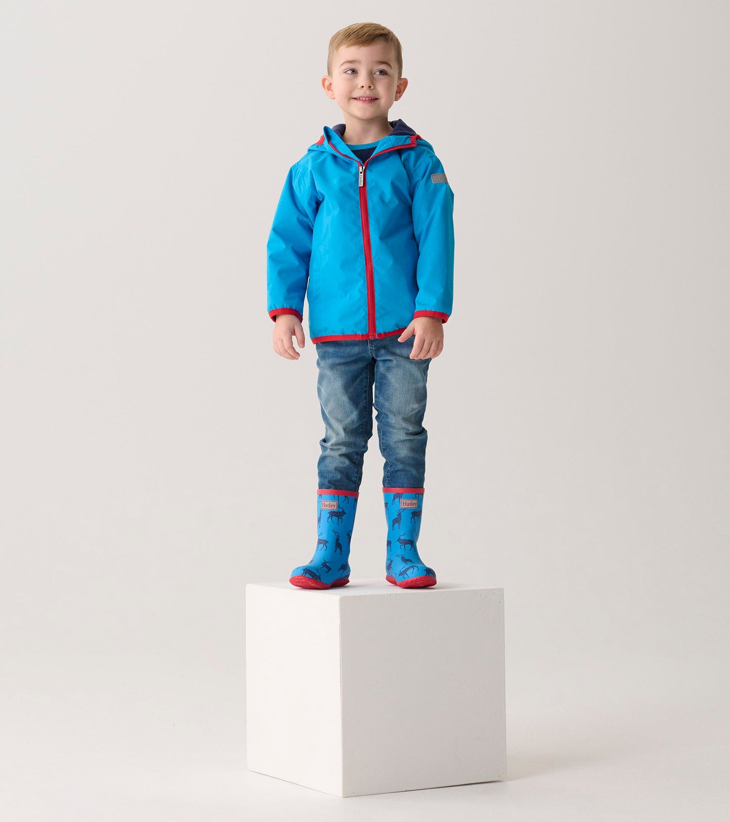 Lightweight blue zip-up rain jacket with red trim, worn by a boy standing on a cube, paired with blue rain boots.