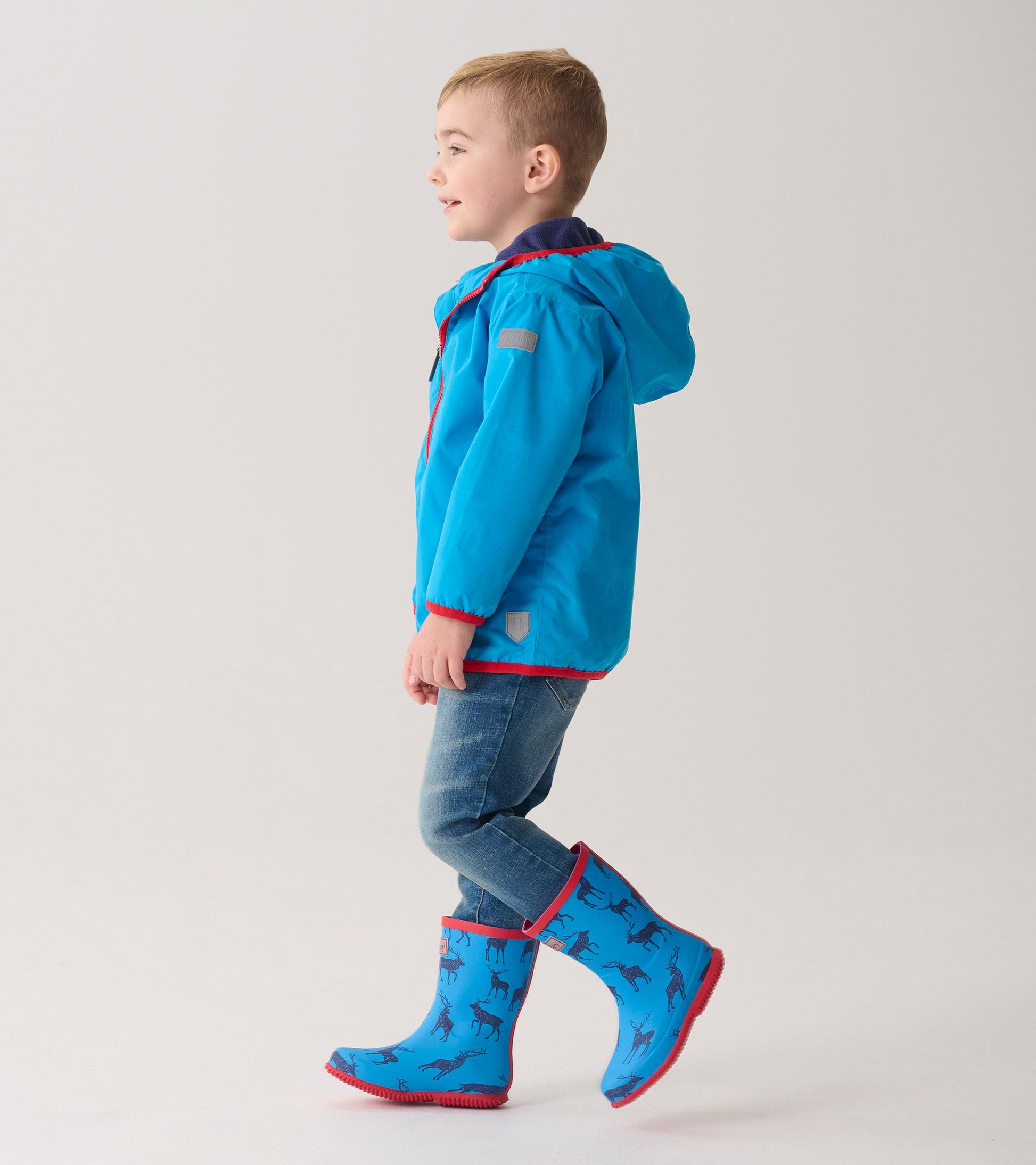 Lightweight blue zip-up rain jacket with a hood, paired with blue boots featuring a horse pattern, worn by a smiling boy.