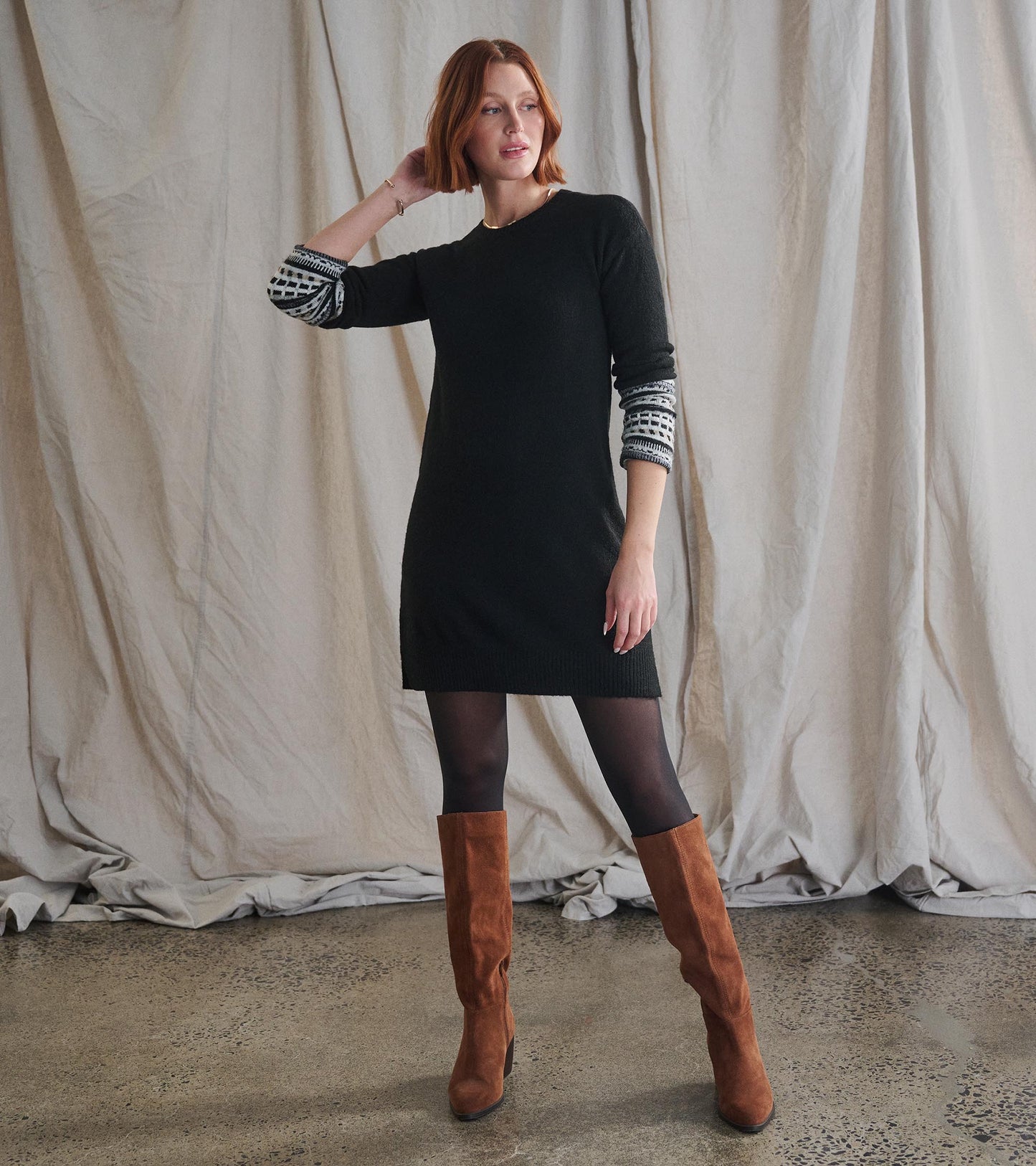 Black sweater dress with long sleeves featuring a patterned cuff, styled with brown knee-high boots.