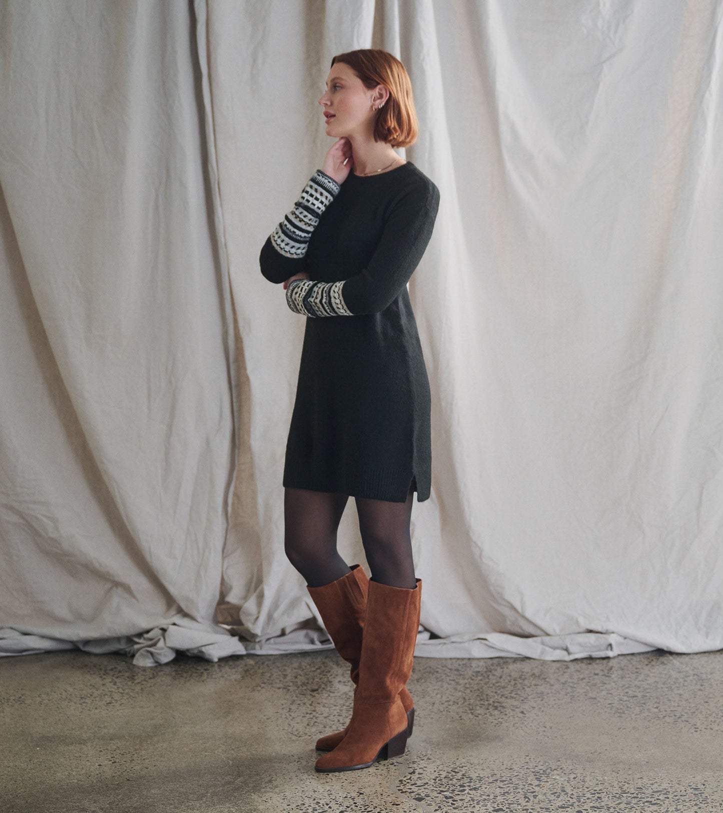 Black sweater dress with patterned sleeves, styled with knee-high brown boots, standing against a neutral backdrop.