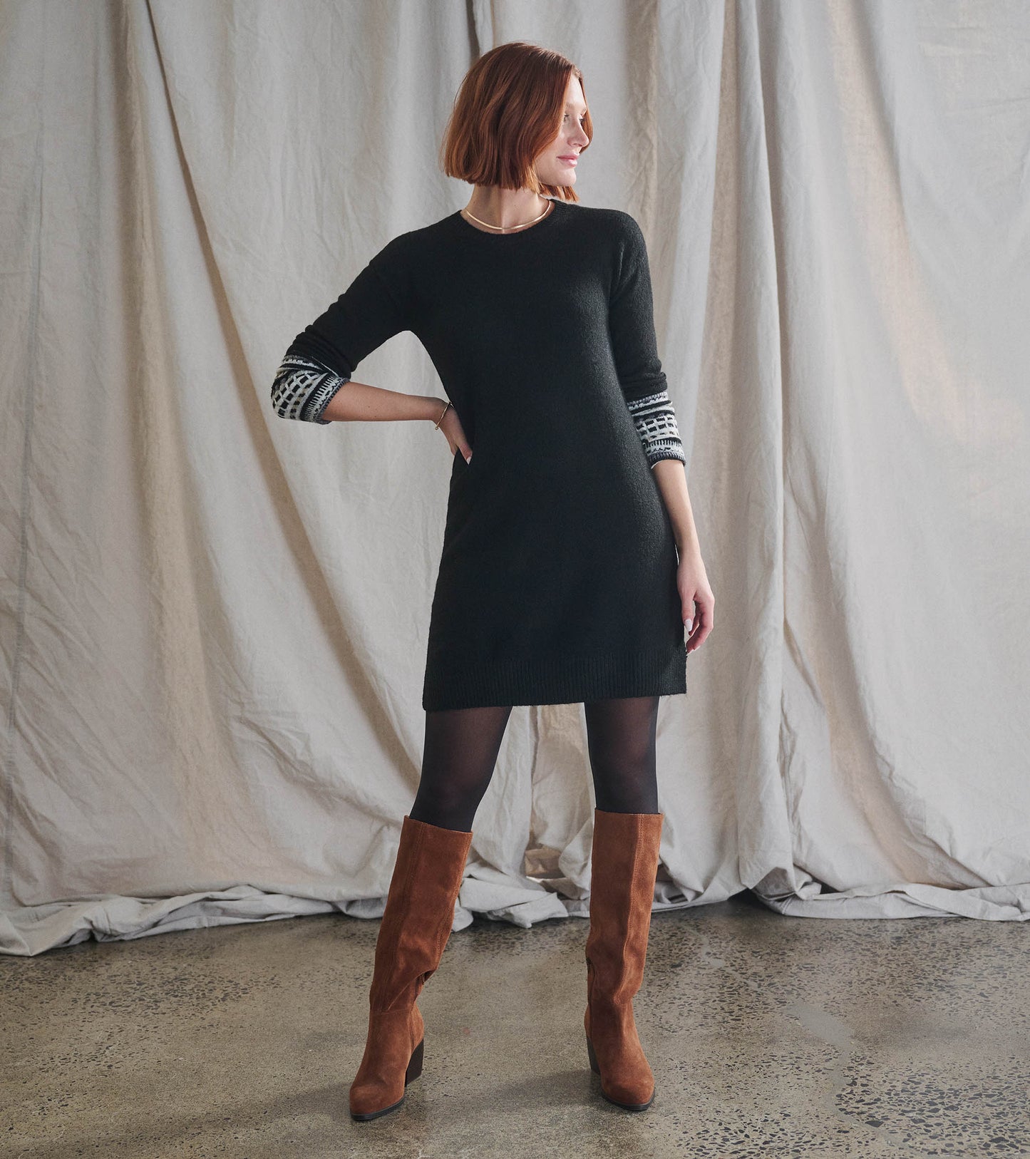 Black sweater dress with long sleeves featuring a patterned cuff, styled with brown knee-high boots.