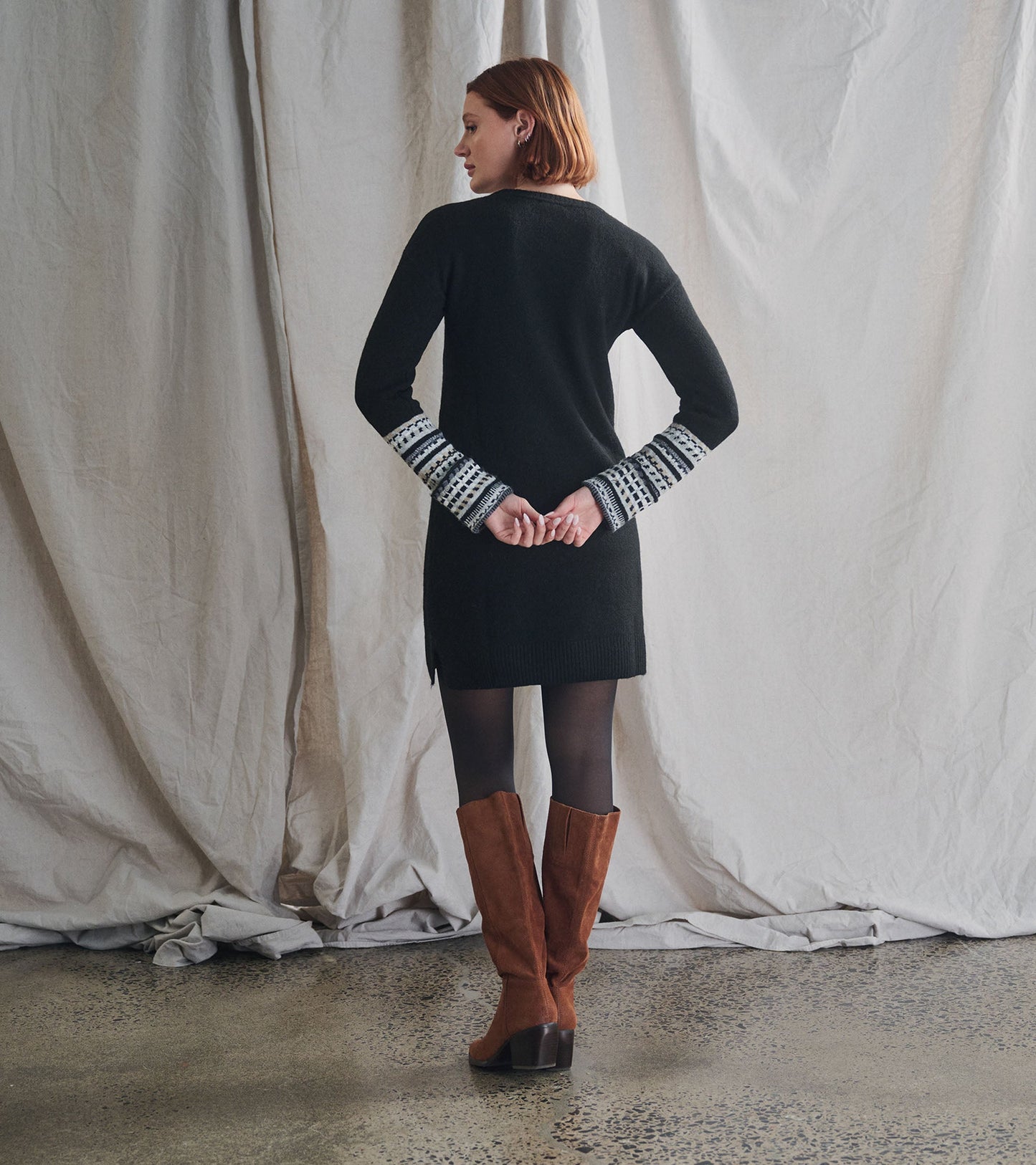 Black sweater dress with patterned sleeves, featuring a relaxed fit and side slits, styled with knee-high brown boots.
