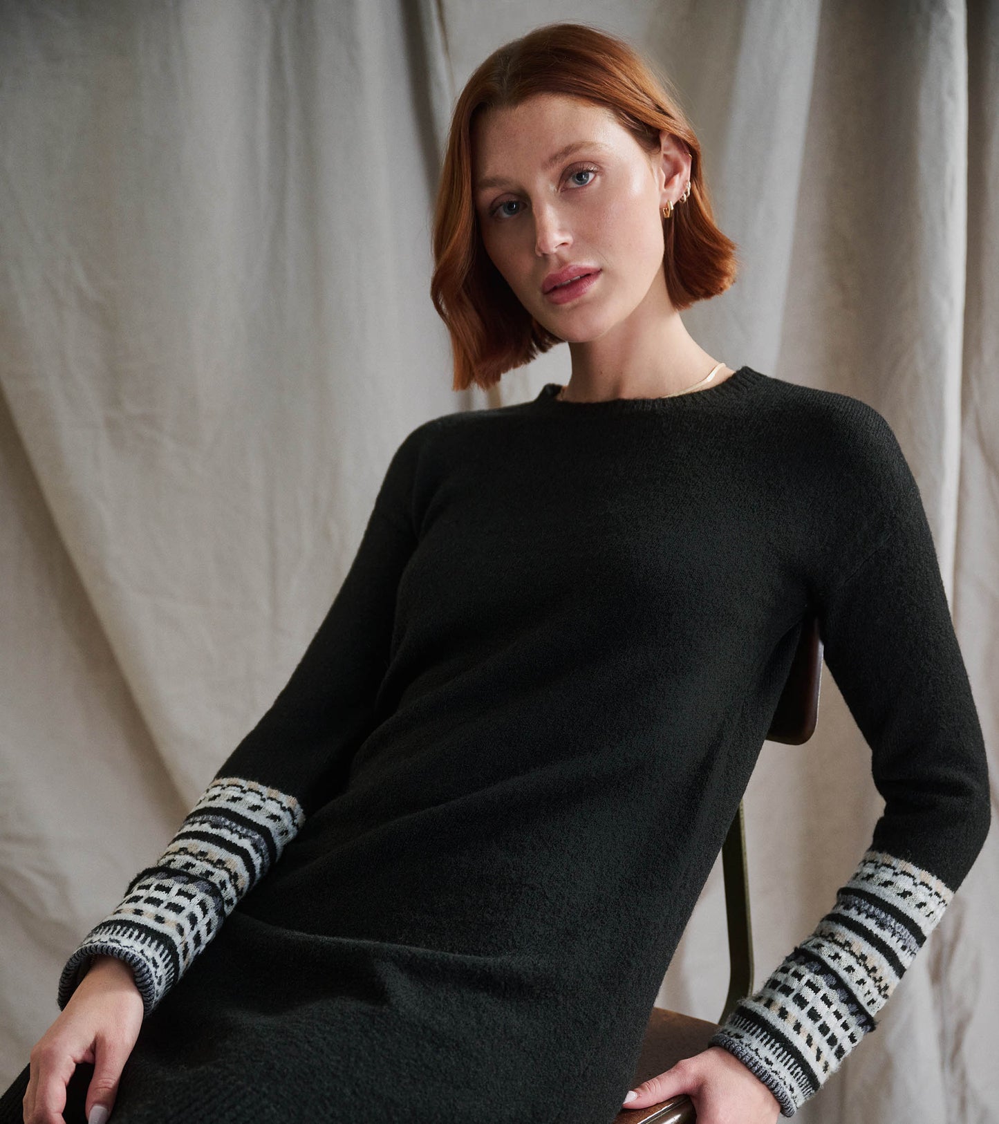 Black sweater dress featuring patterned cuffs, long sleeves, and a relaxed fit, styled with a model seated.