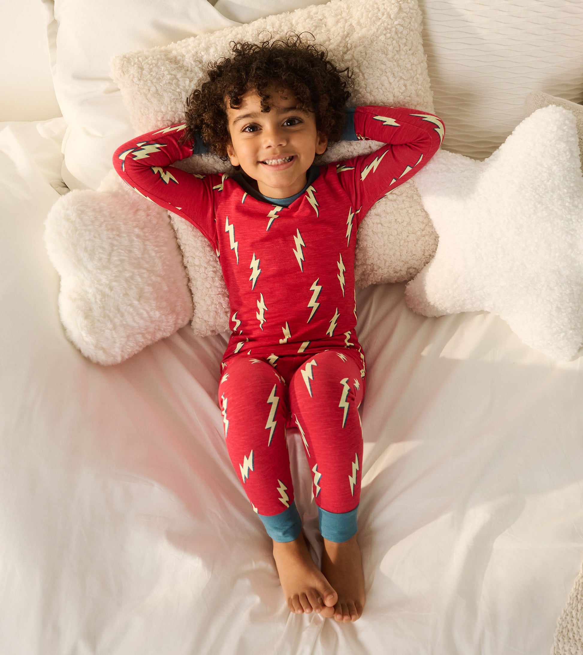 Red pajama set featuring lightning bolt pattern, with blue cuffs, worn by a child lounging on a bed with fluffy pillows.