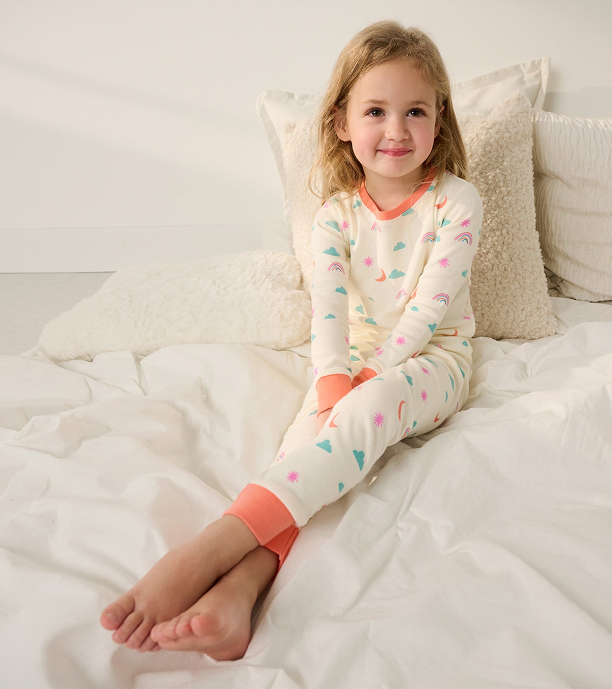 Organic cotton pajama set in cream with colorful cloud, rainbow, and star patterns, featuring coral cuffs and neckline.
