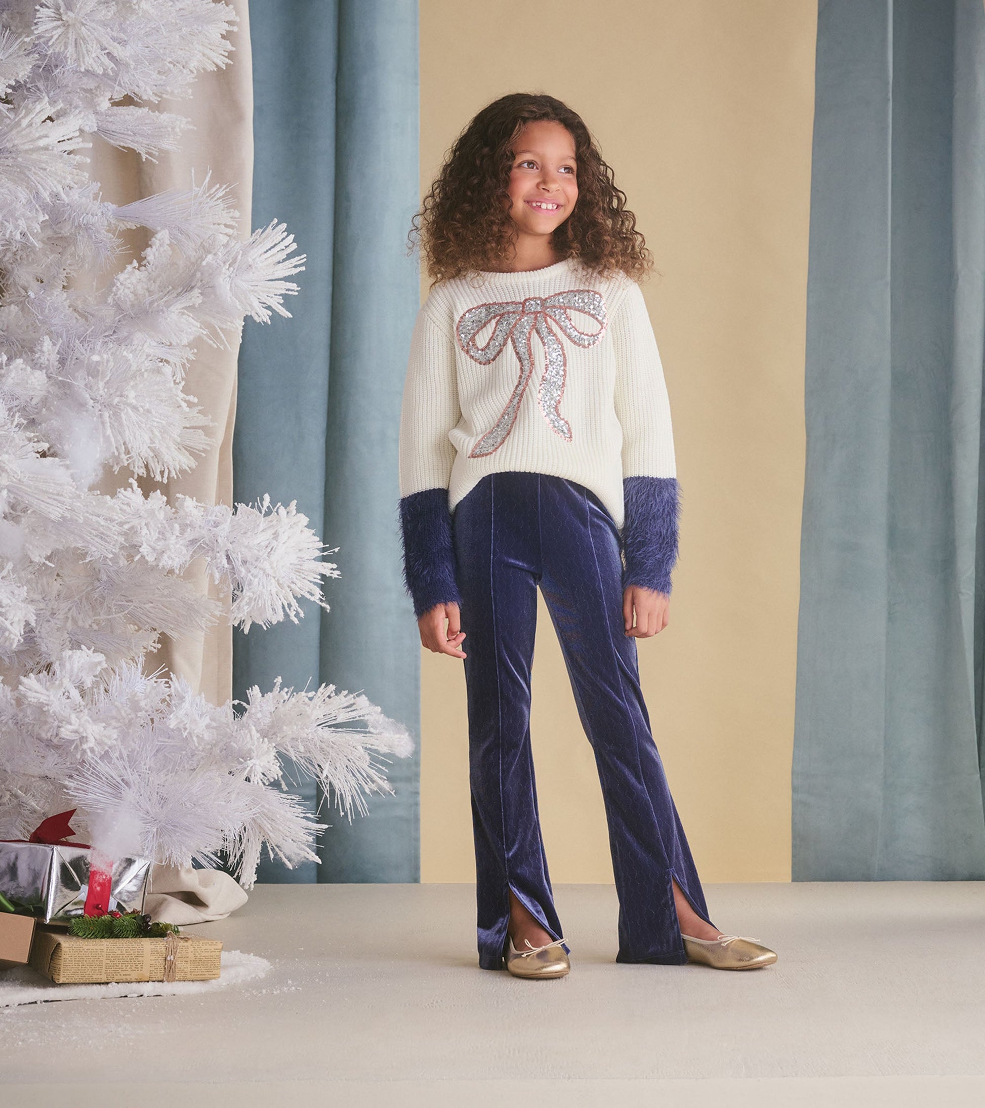 Cream sweater with sequin bow detail and fluffy blue sleeves, paired with navy velvet pants, worn by a smiling girl.