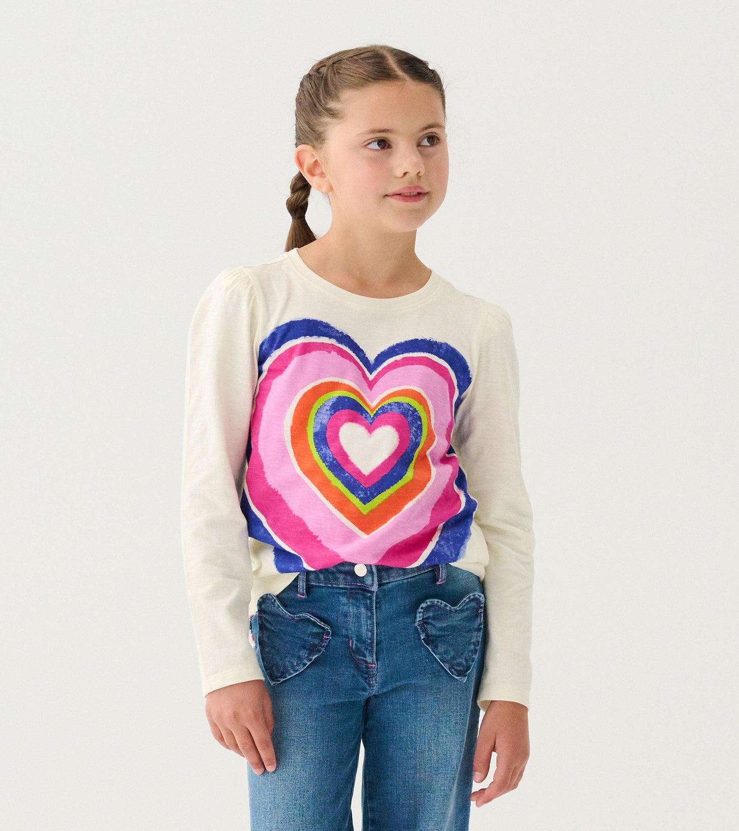 Long sleeve cream graphic tee featuring a colorful layered heart design, paired with jeans with heart-shaped pockets.