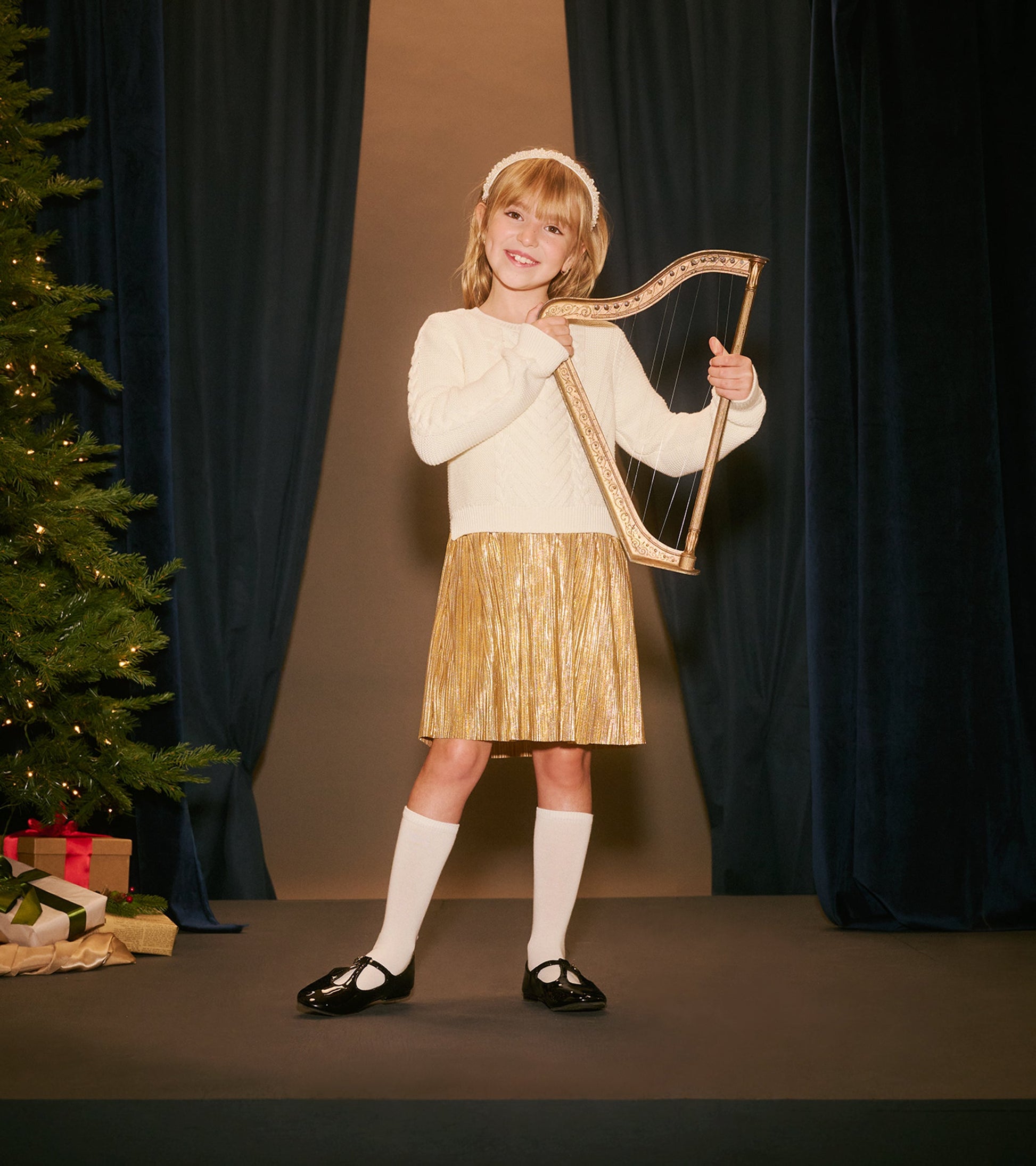 Cream knit top with a gold pleated skirt, worn by a girl holding a harp, set against a festive backdrop.