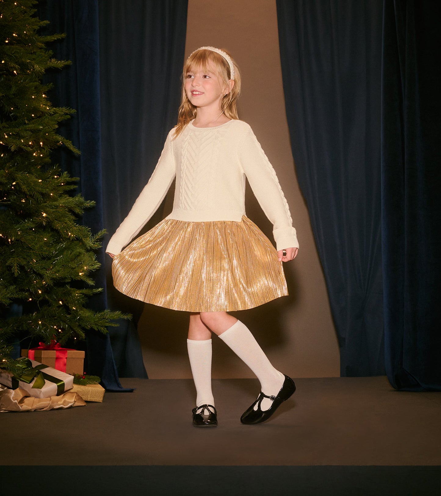Cream long-sleeve top with cable knit design paired with a shiny gold pleated skirt, worn with white socks and black shoes.