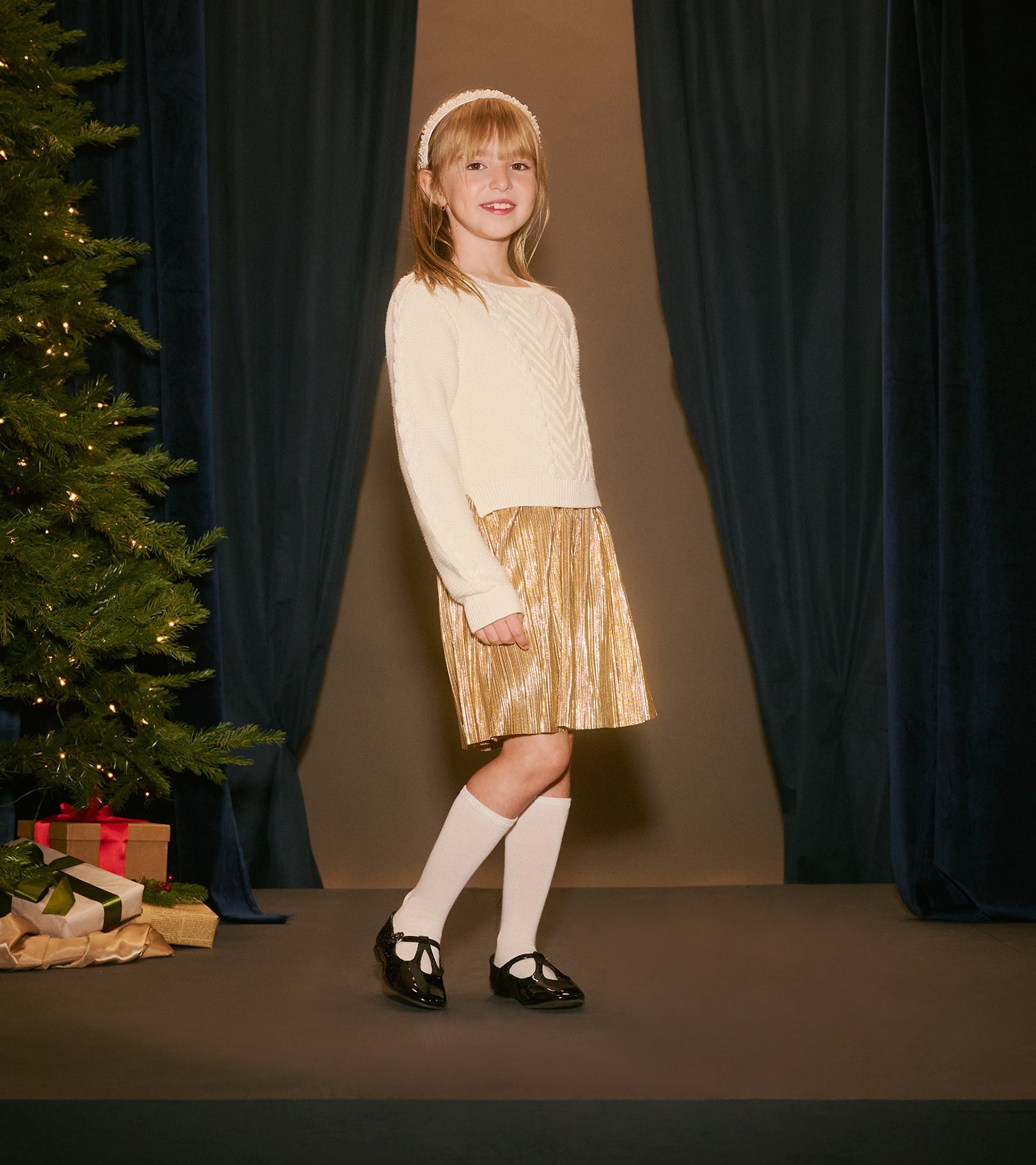 Cream sweater with a textured pattern paired with a shiny gold pleated skirt, worn with knee-high socks and black shoes.