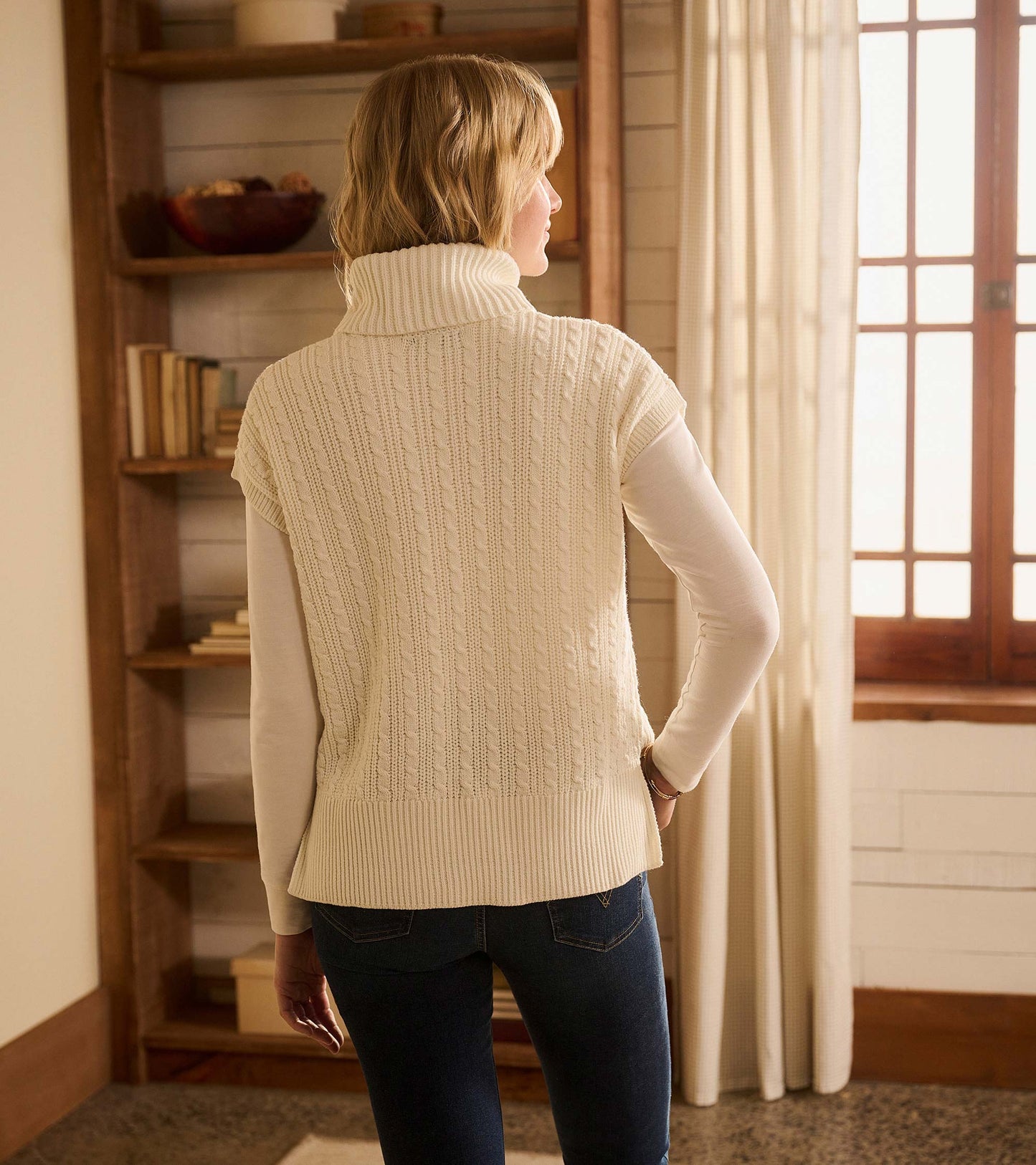 Cable knit vest in winter white with a high collar, worn over a long-sleeve top, viewed from the back.