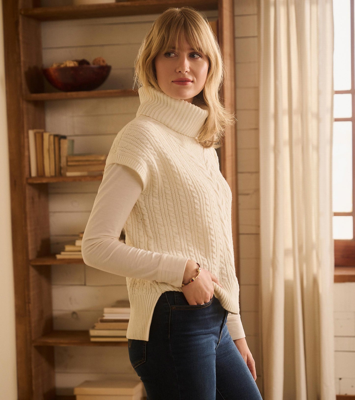 Cable knit vest in winter white with a high collar, worn over a long-sleeve top, styled with jeans.