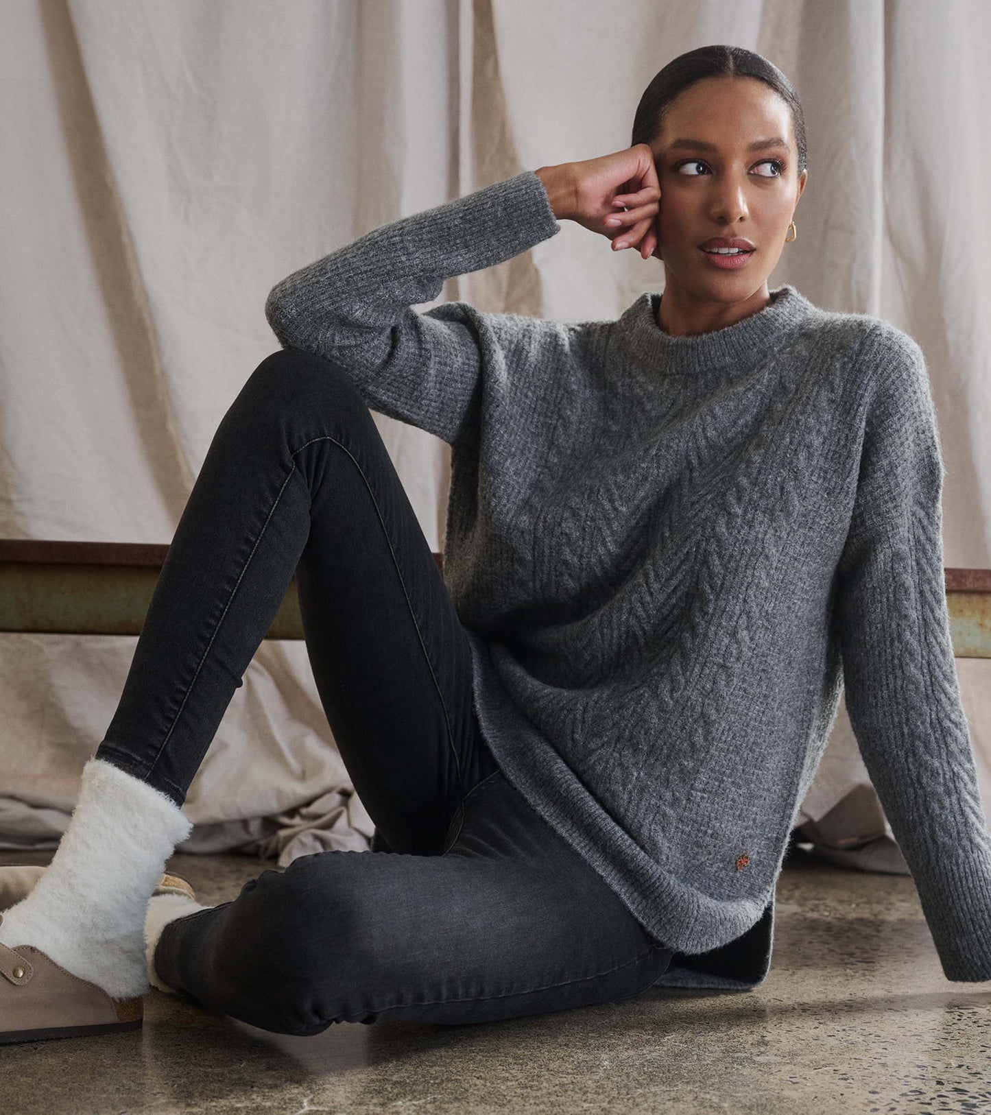Charcoal heather cable knit tunic with a relaxed fit, paired with black jeans and cozy socks, seated against a backdrop.