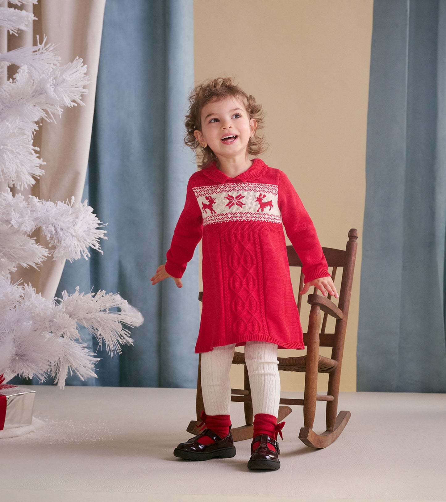 Red cable knit sweater dress with festive reindeer pattern, paired with white tights and black shoes, worn by a smiling chil…