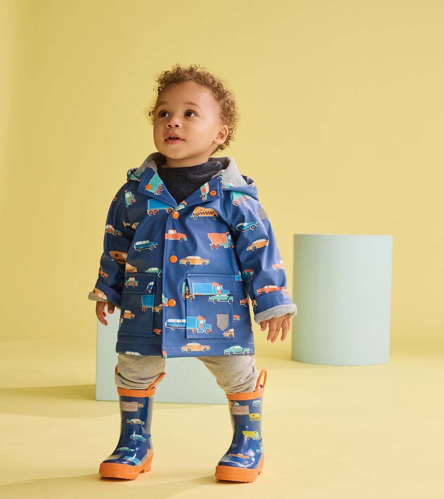 Blue rain jacket featuring colorful driving cars, with a hood and front pockets, worn by a toddler in a playful setting.
