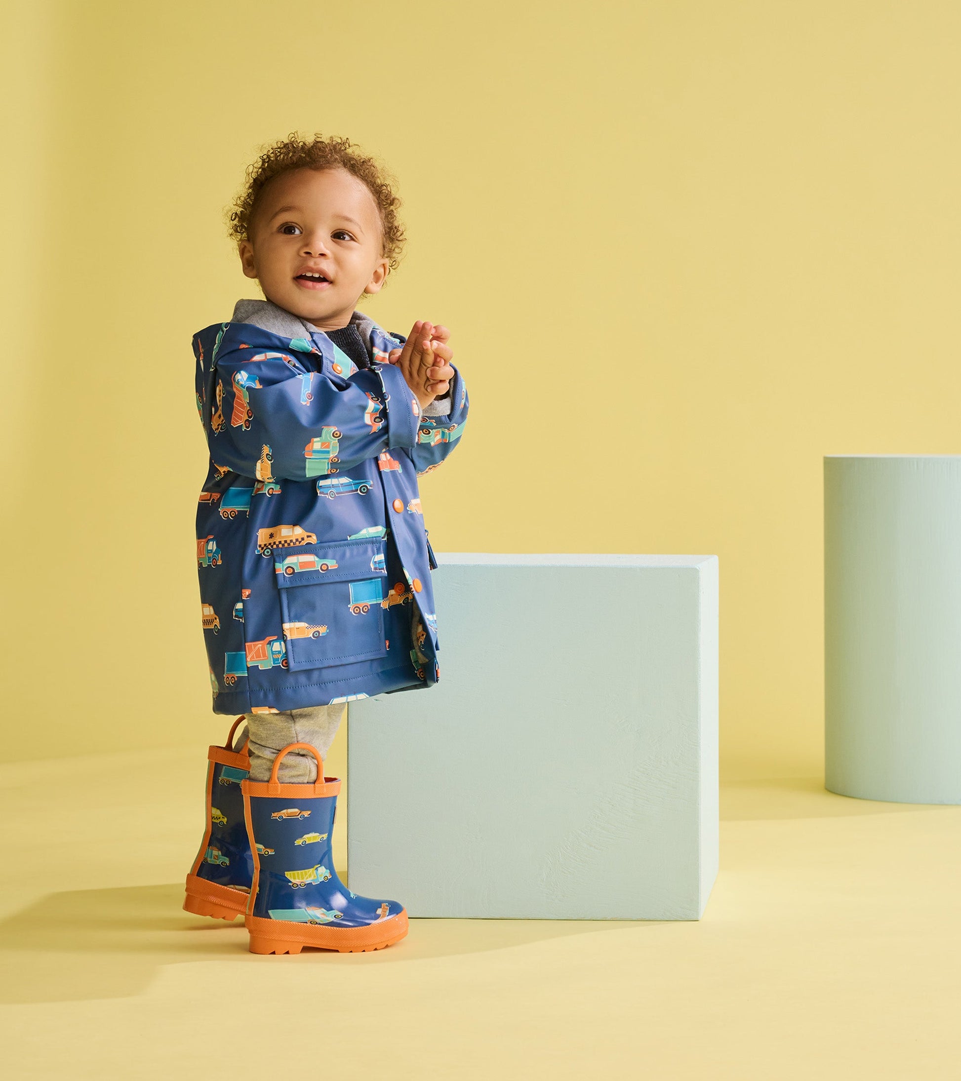 Blue rain jacket with colorful car patterns, worn by a toddler, paired with matching blue and orange rain boots.