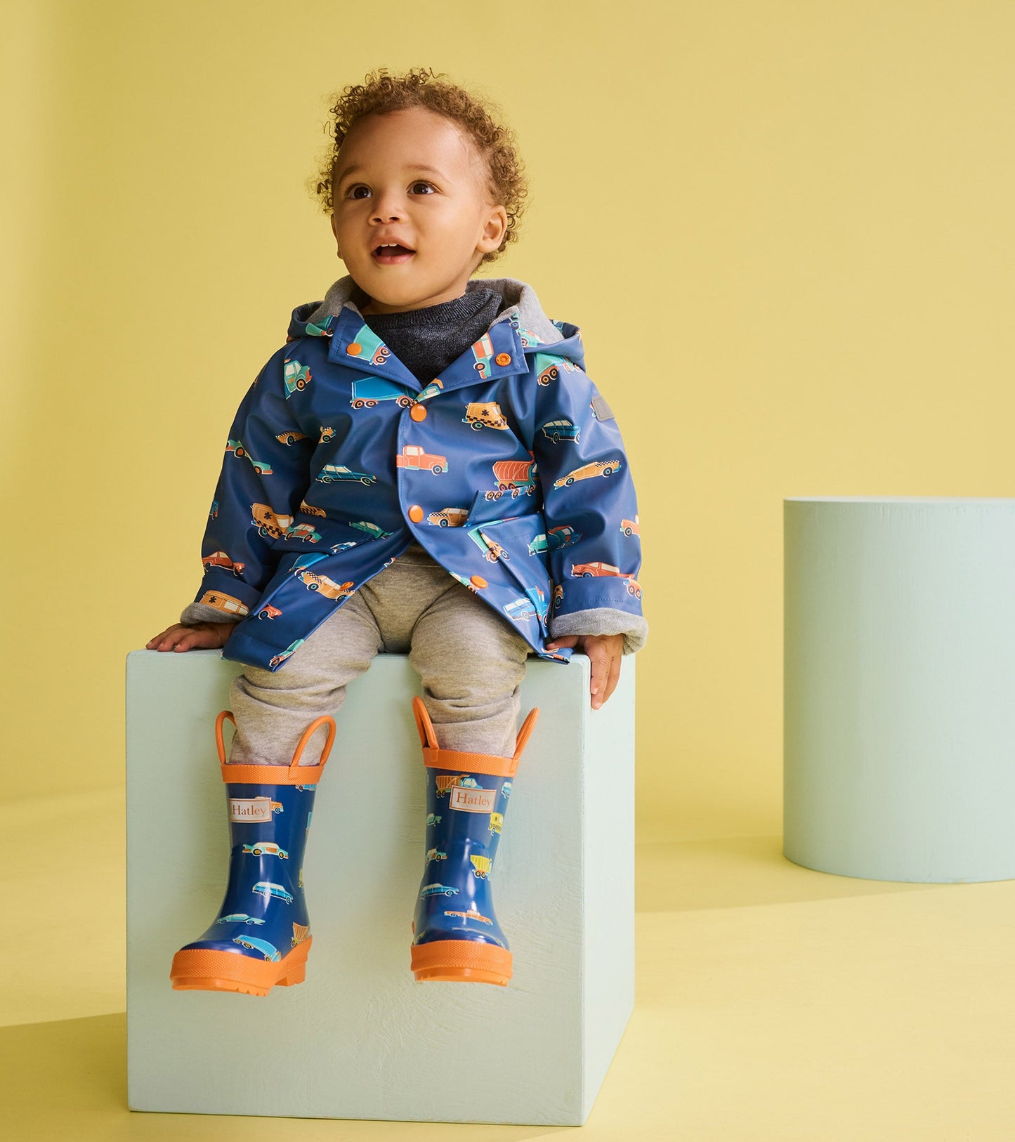 Blue rain jacket with colorful car patterns, worn by a toddler, paired with gray pants and matching rain boots.