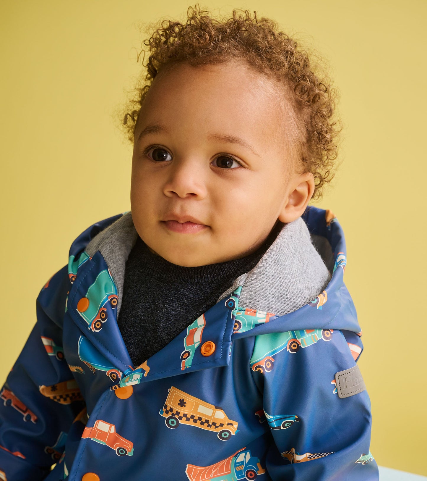 Blue rain jacket featuring colorful driving cars pattern, with a gray hood and orange buttons, suitable for toddlers.