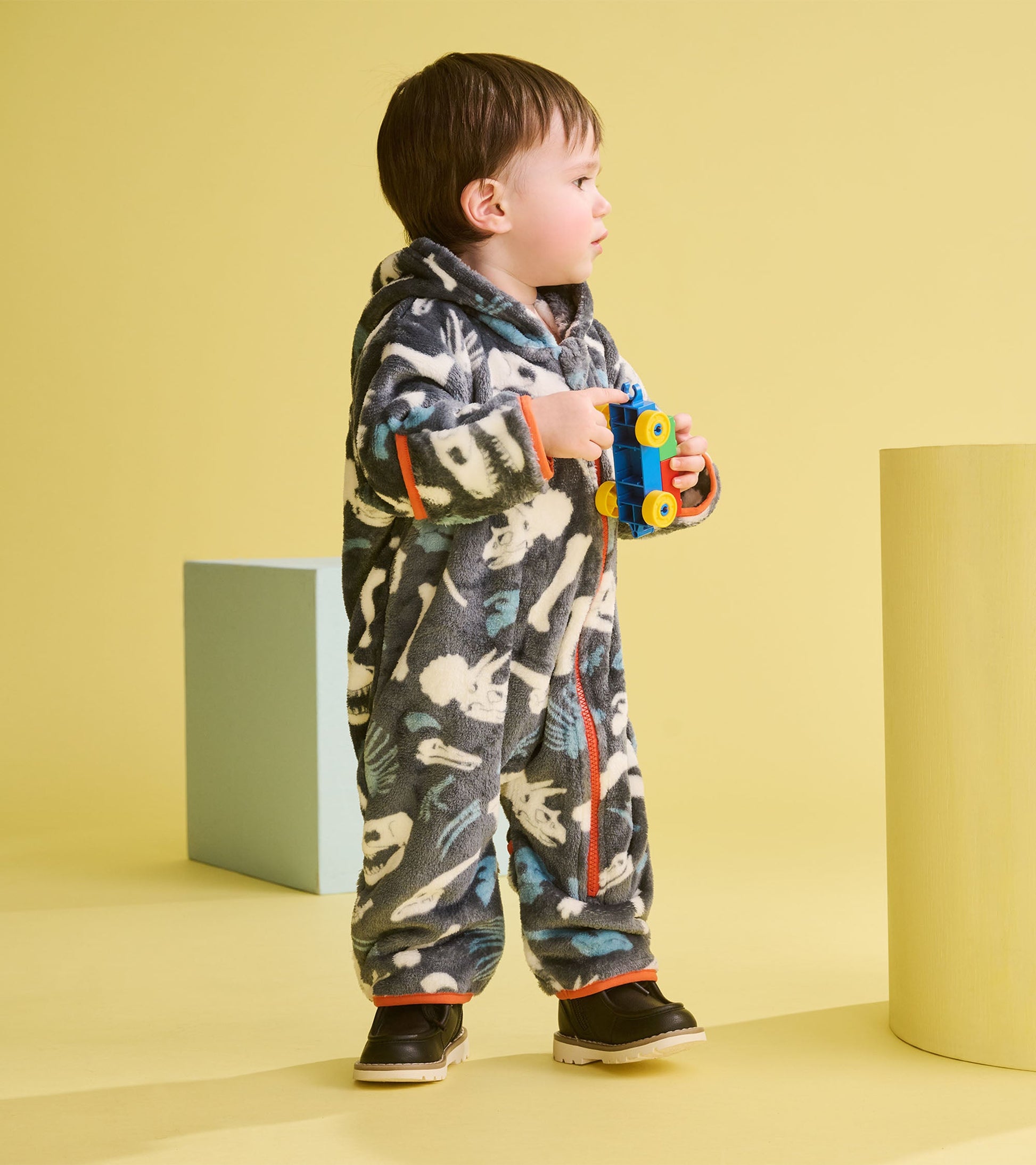Gray fleece suit with dinosaur fossil patterns, featuring a hood and orange accents, worn by a child holding a toy.