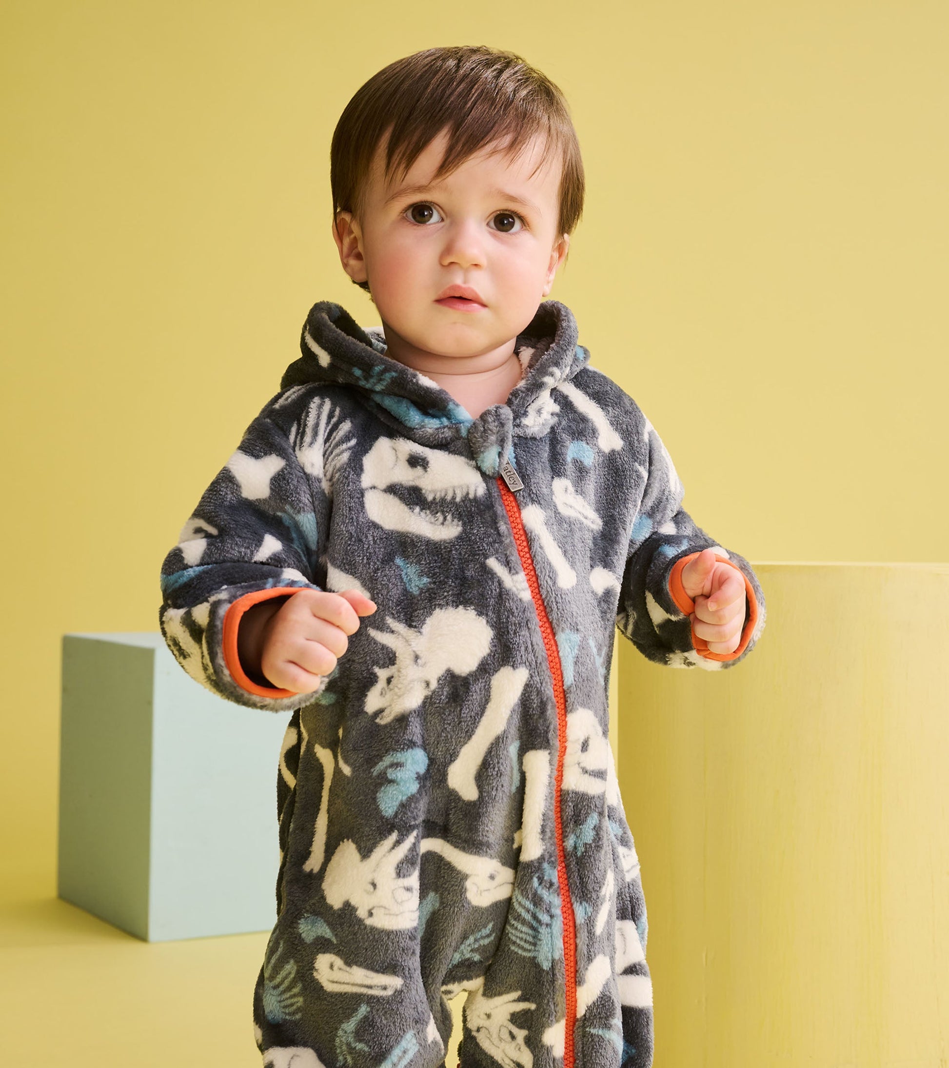 Gray fleece suit with dinosaur fossil print, featuring a hood and orange zipper, worn by a baby boy.