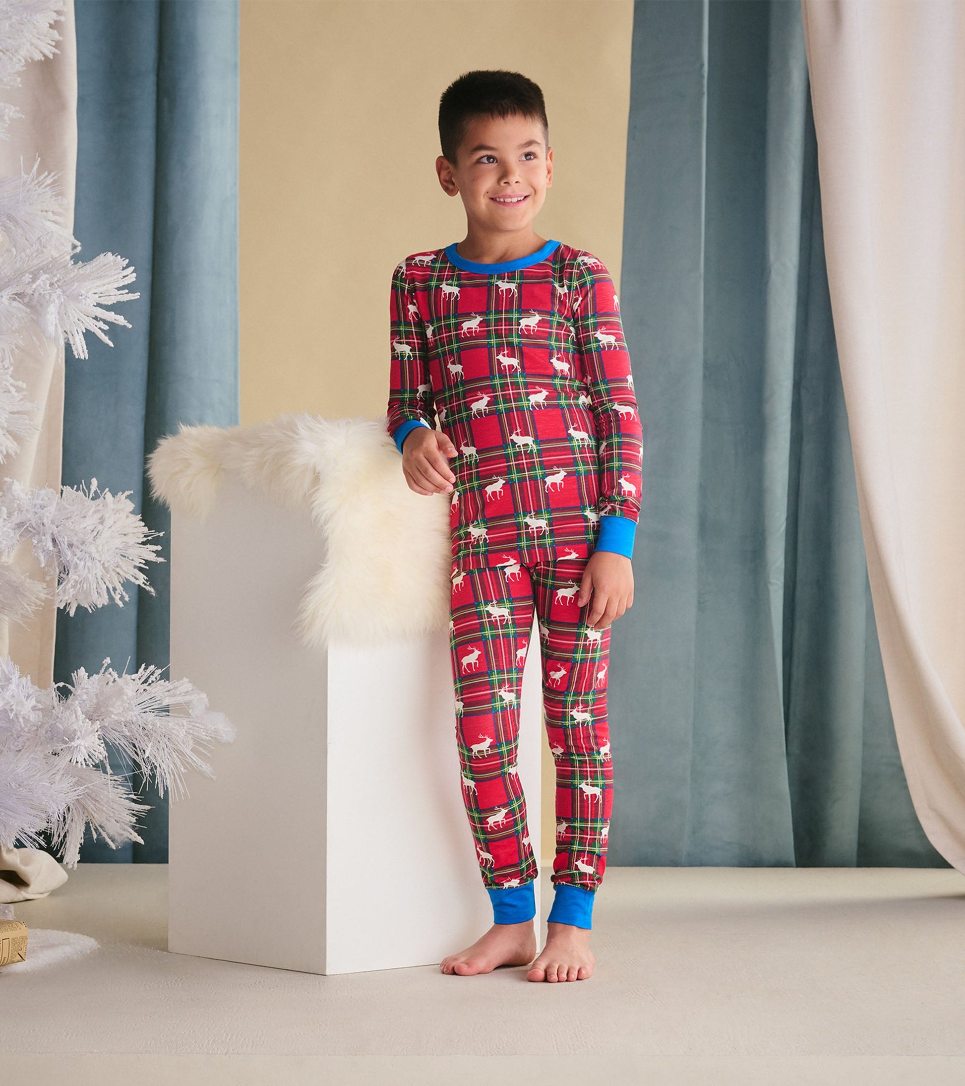 Red plaid pajama set featuring elk patterns, with blue cuffs and neckline, worn by a smiling boy.