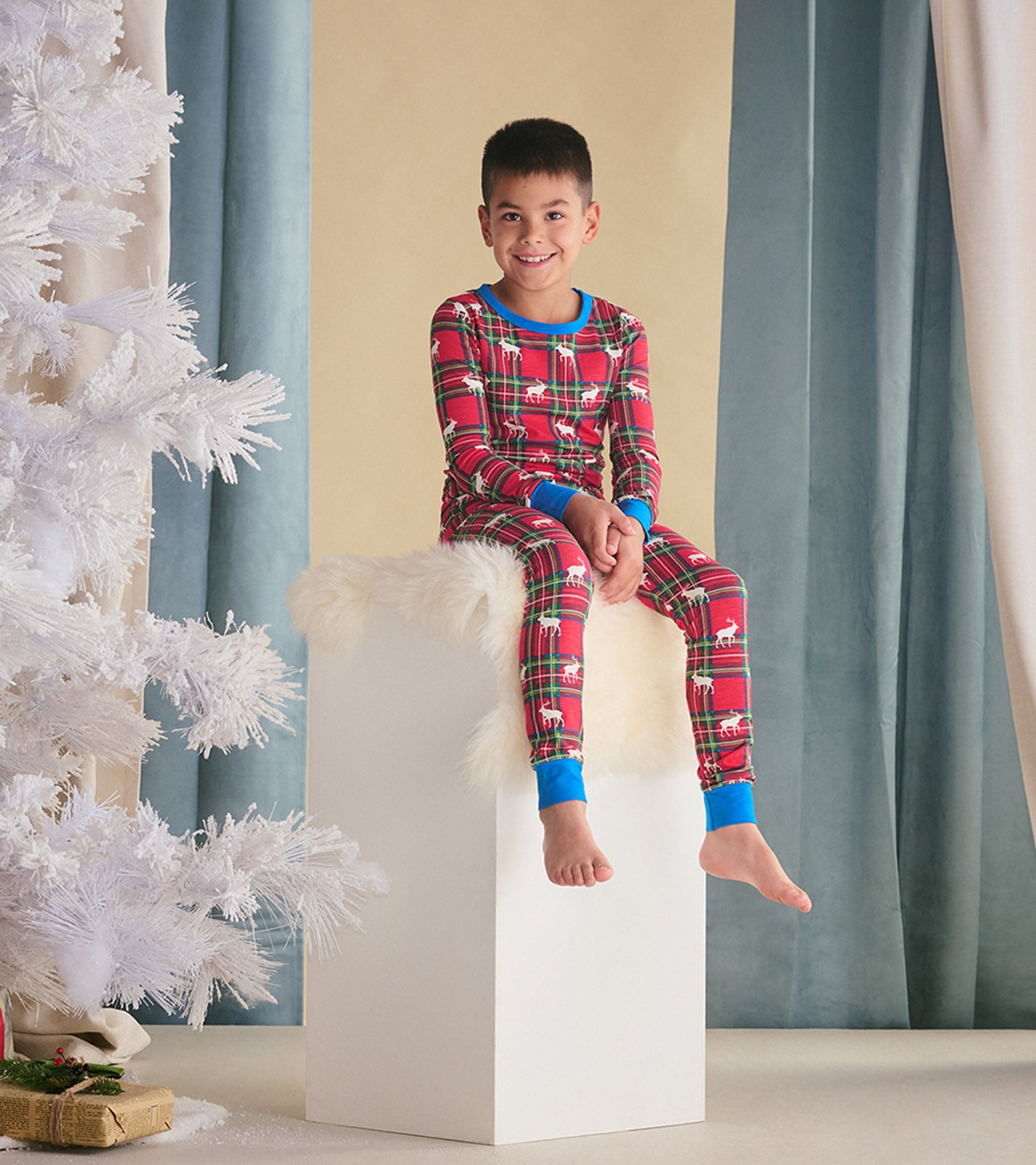 Red plaid pajama set with elk print, featuring blue cuffs, worn by a smiling boy sitting on a white pedestal.