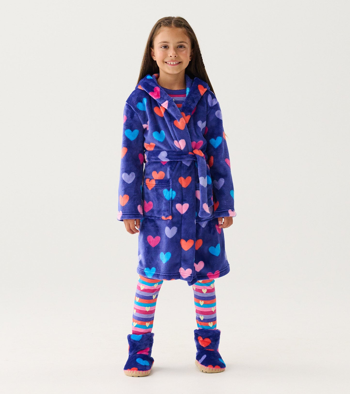 Purple fleece robe with colorful heart patterns, featuring a hood, belt, and pockets, worn over striped leggings and slipper…