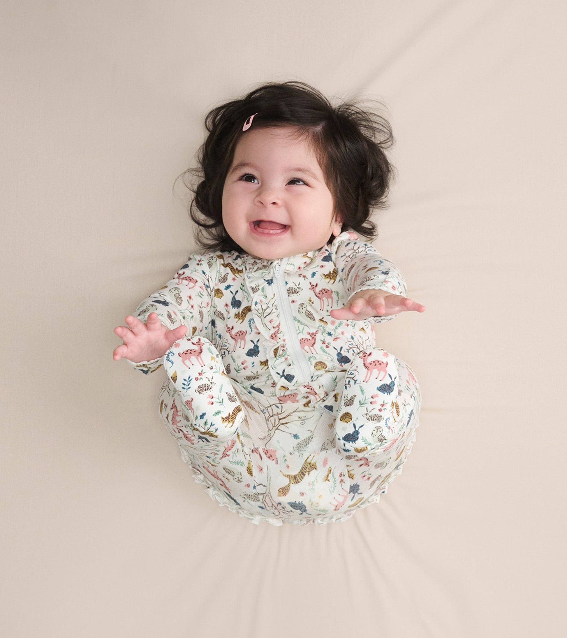 Baby girl in a ruffle bum bamboo sleeper with a colorful forest animal print, smiling while lying on a light background.