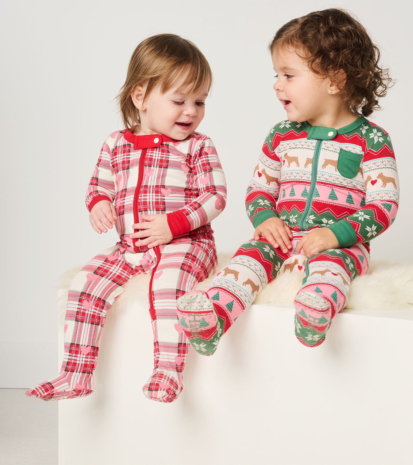 Two toddlers sit side by side wearing colorful cotton footed sleepers with plaid and festive patterns.