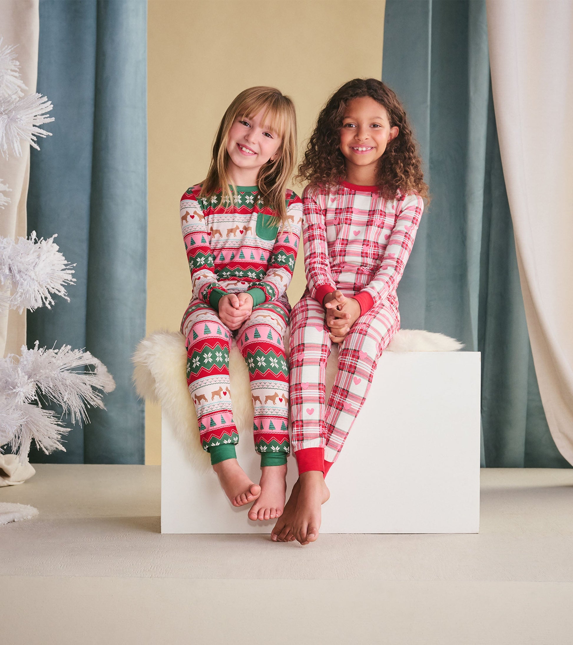 Two girls sit side by side wearing colorful plaid and patterned pajamas, smiling, with bare feet on a white platform.