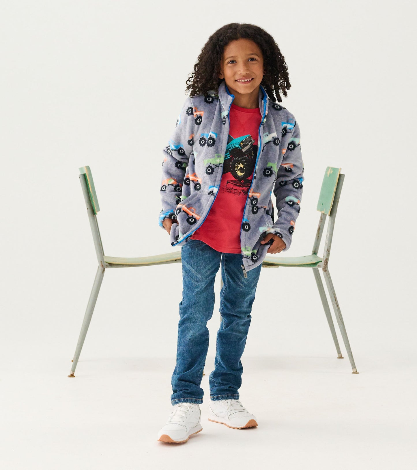 Fuzzy fleece zip-up jacket featuring colorful monster truck patterns, paired with a red shirt and blue jeans.