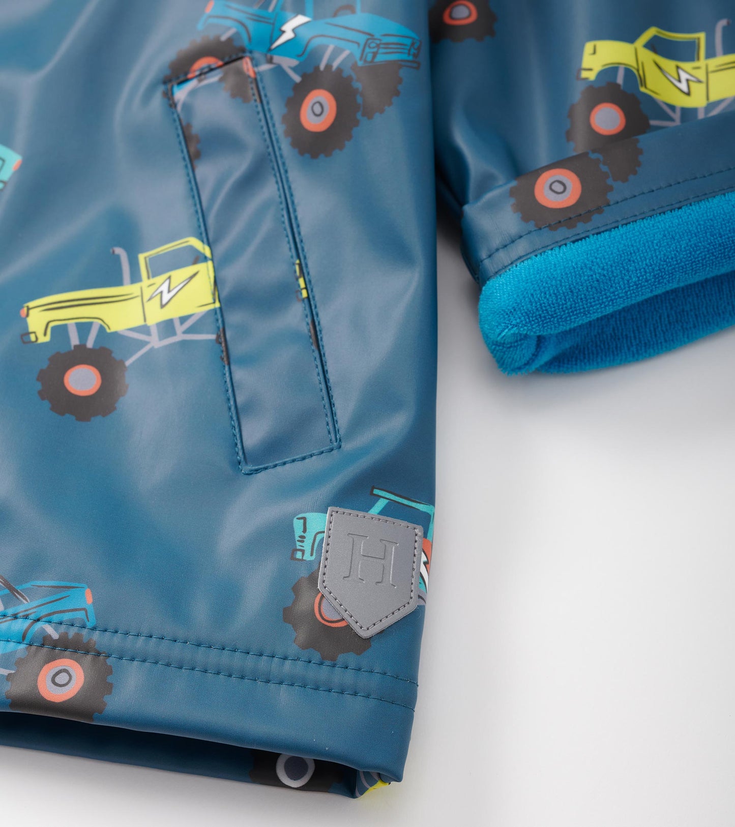 Blue zip-up rain jacket featuring colorful monster truck patterns and a soft blue fleece lining at the cuffs.