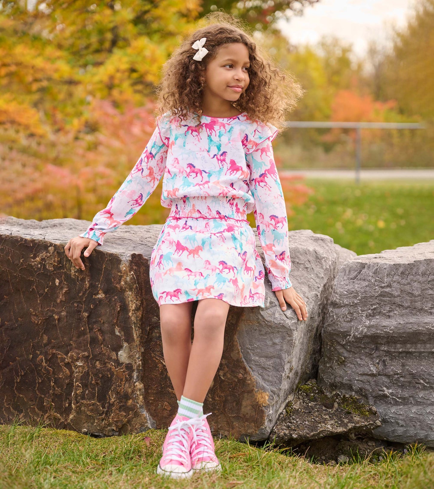 Light pink dress with colorful horse print, smocked waist, long sleeves, and ruffled neckline, worn with pink sneakers.