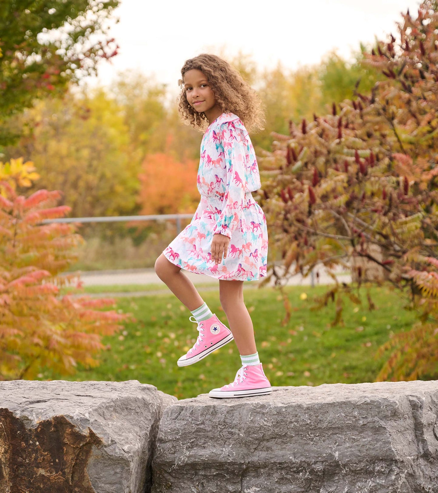 Light pink dress with colorful horse print and smocked waist, paired with pink sneakers and green socks, worn outdoors.