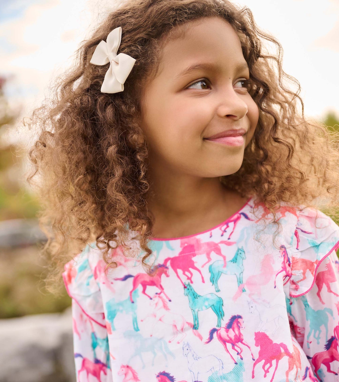 Colorful dress featuring a smocked waist and playful horse print, worn by a girl with curly hair and a bow.