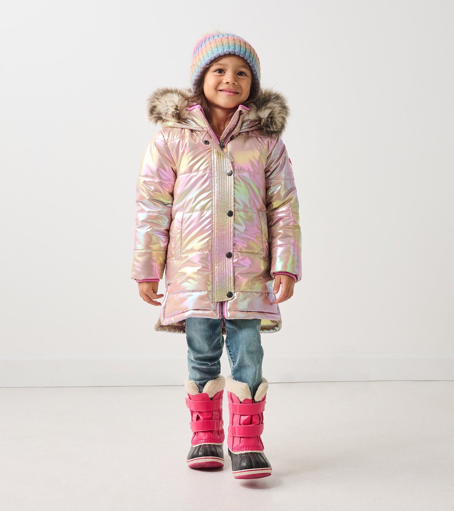 Iridescent pink puffer jacket with faux fur hood, paired with blue jeans and pink winter boots.