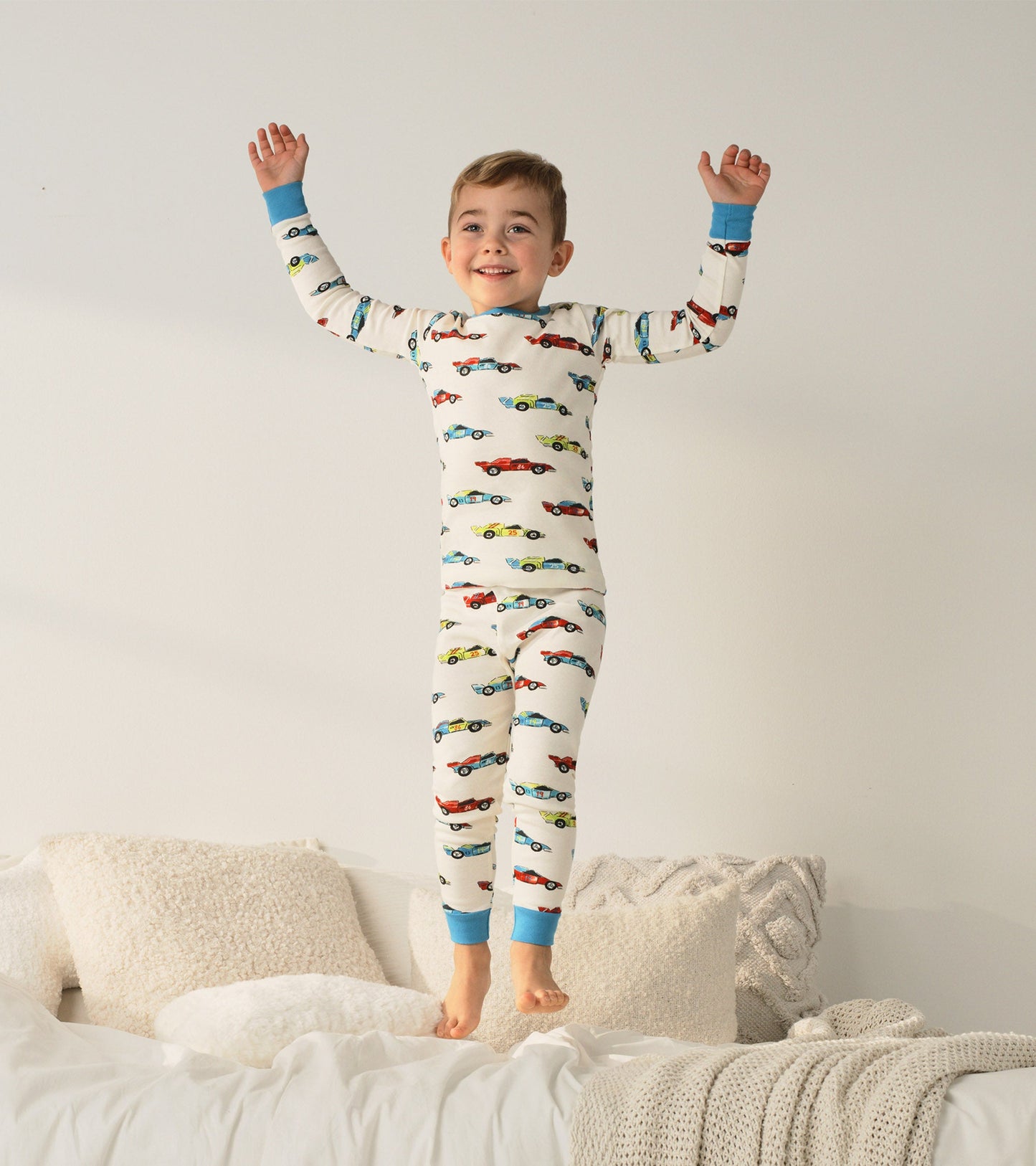 Organic cotton pajama set featuring colorful racecars on a cream background, with blue cuffs, worn by a smiling boy.