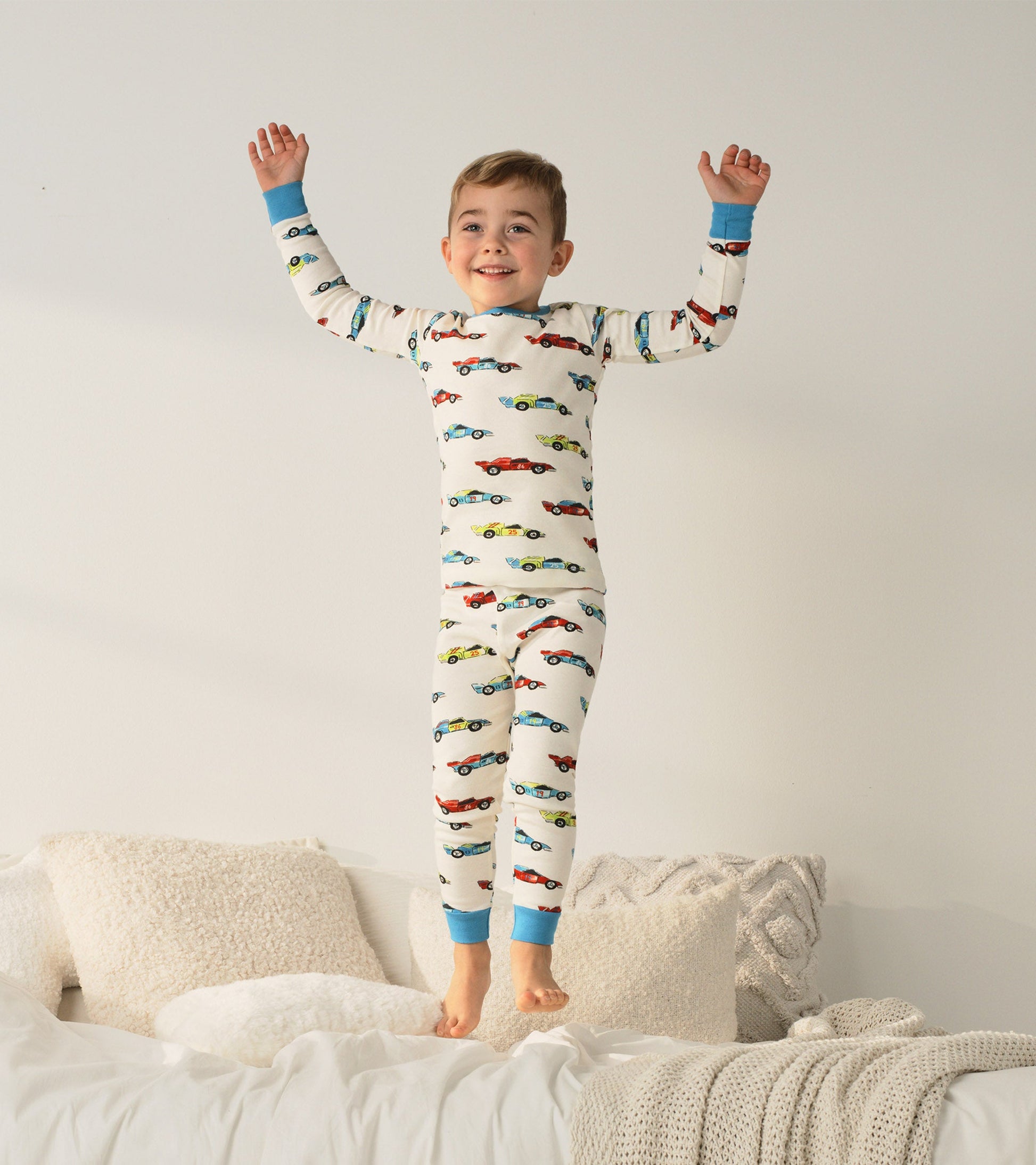 Organic cotton pajama set featuring colorful racecars on a cream background, with blue cuffs, worn by a smiling boy.